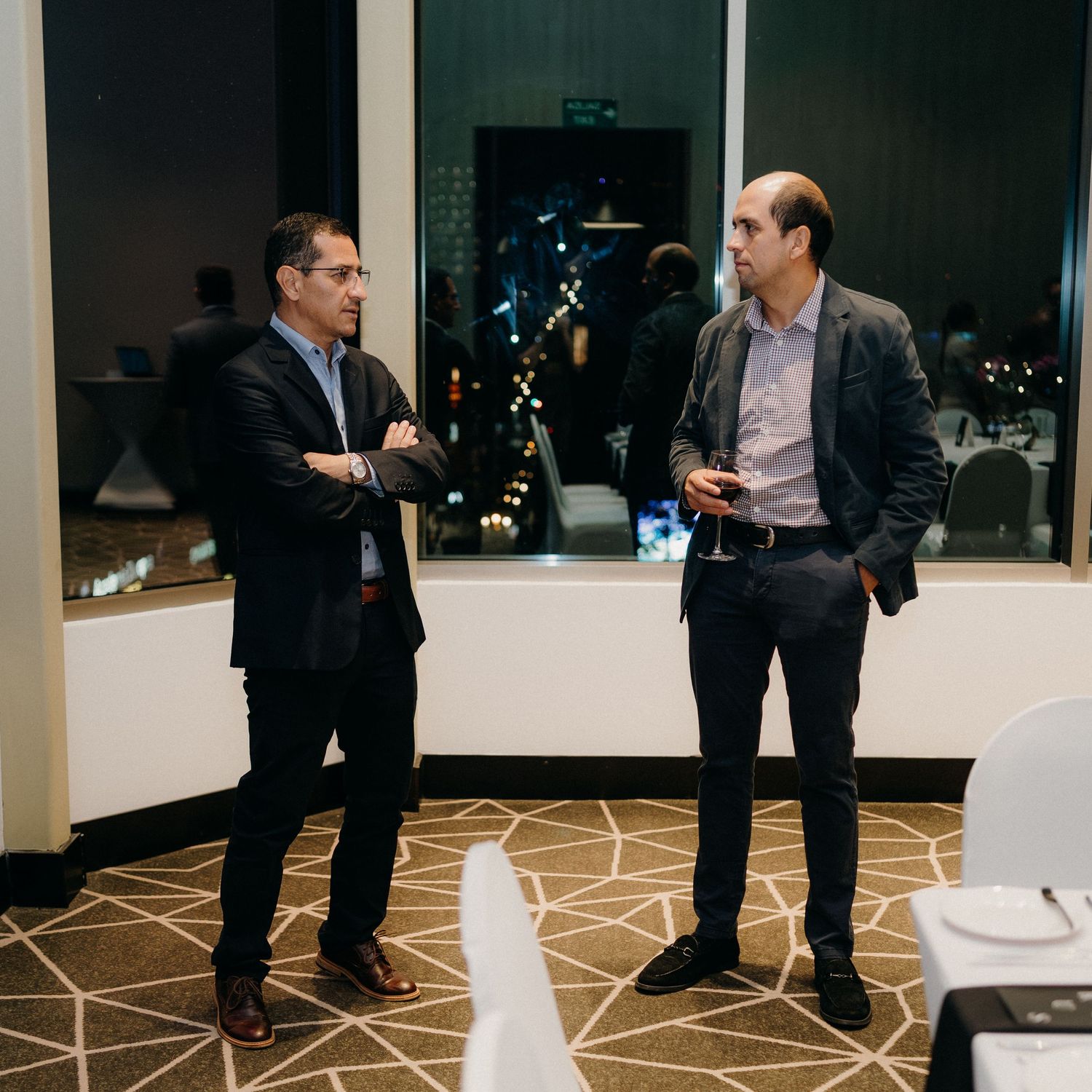 Business professionals in dark suits having conversations in a modern office hallway with geometric patterned carpet.