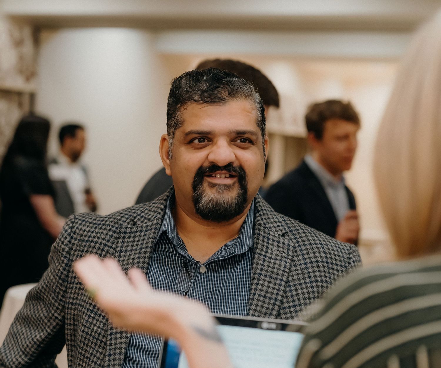 A person in a checkered shirt smiles while holding papers during what appears to be a business meeting or conference.
