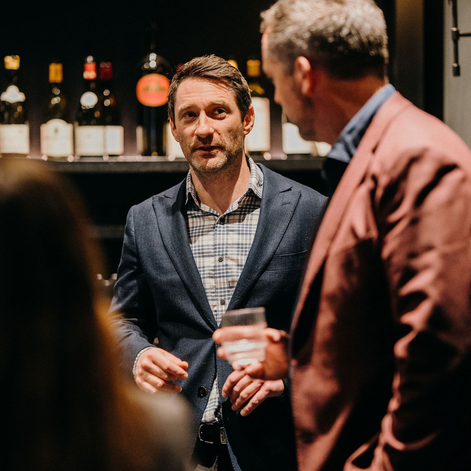 People having drinks and socializing at a dimly lit bar with bottles visible in the background.
