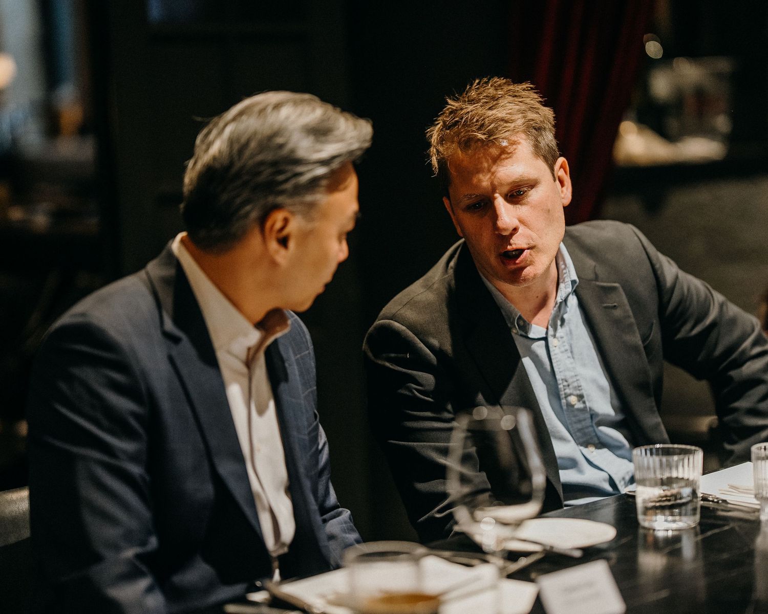 Two men in business attire having an intense discussion at a restaurant table with drinks.