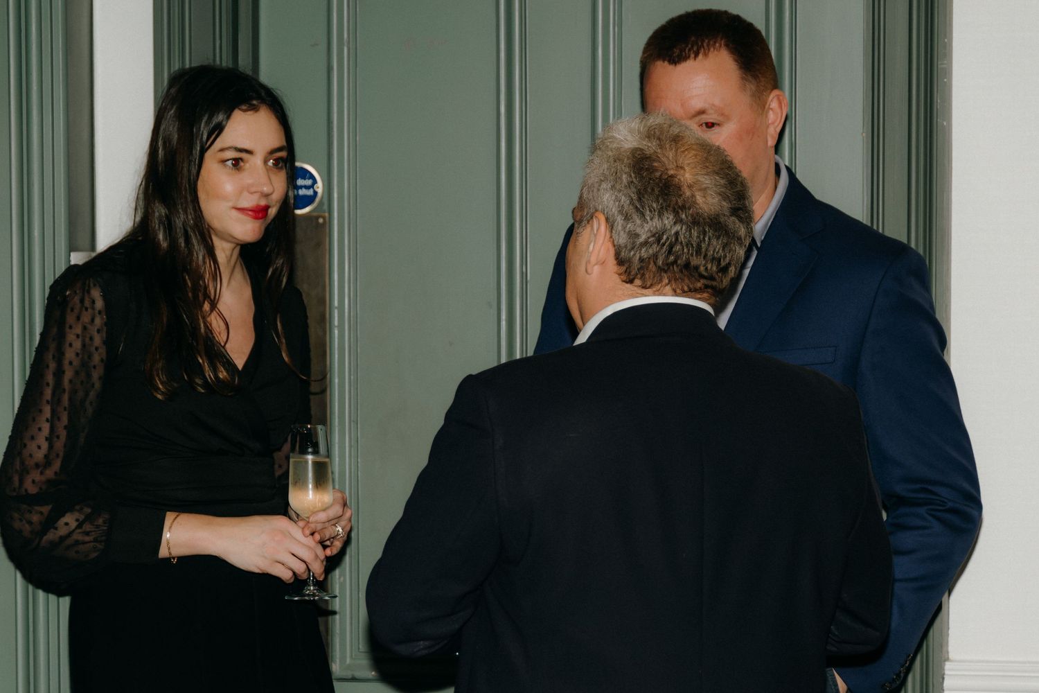People in formal attire gather at an elegant event with green paneled walls in the background.