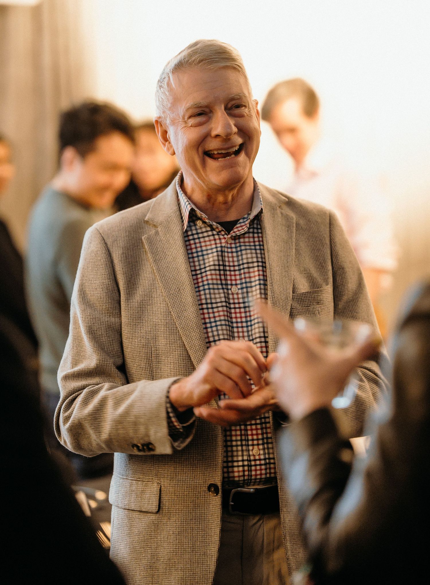 A person wearing a gray blazer and checkered shirt laughs joyfully at a social gathering.