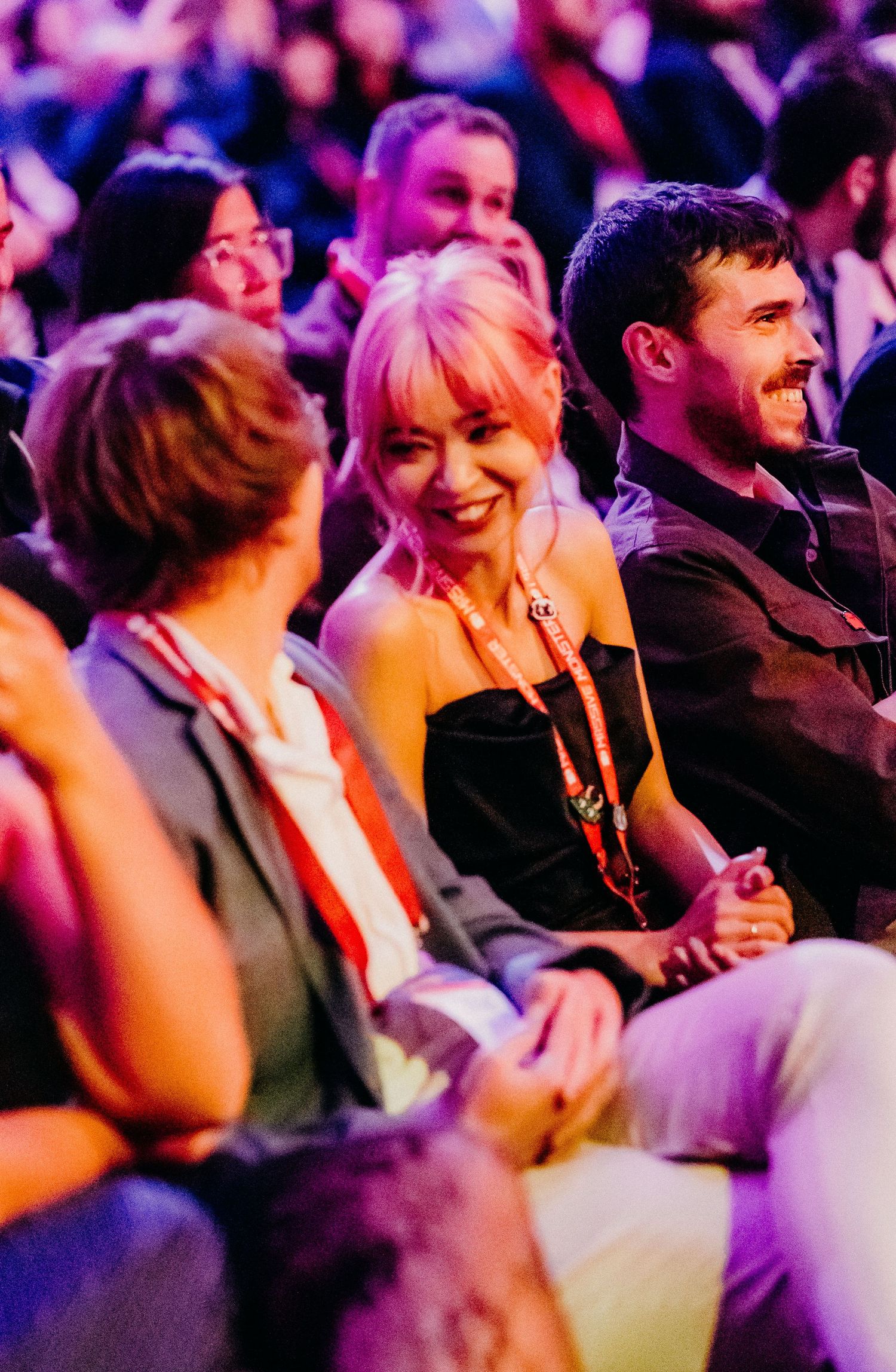 Audience members laugh and smile during a comedy show performance under purple stage lighting.