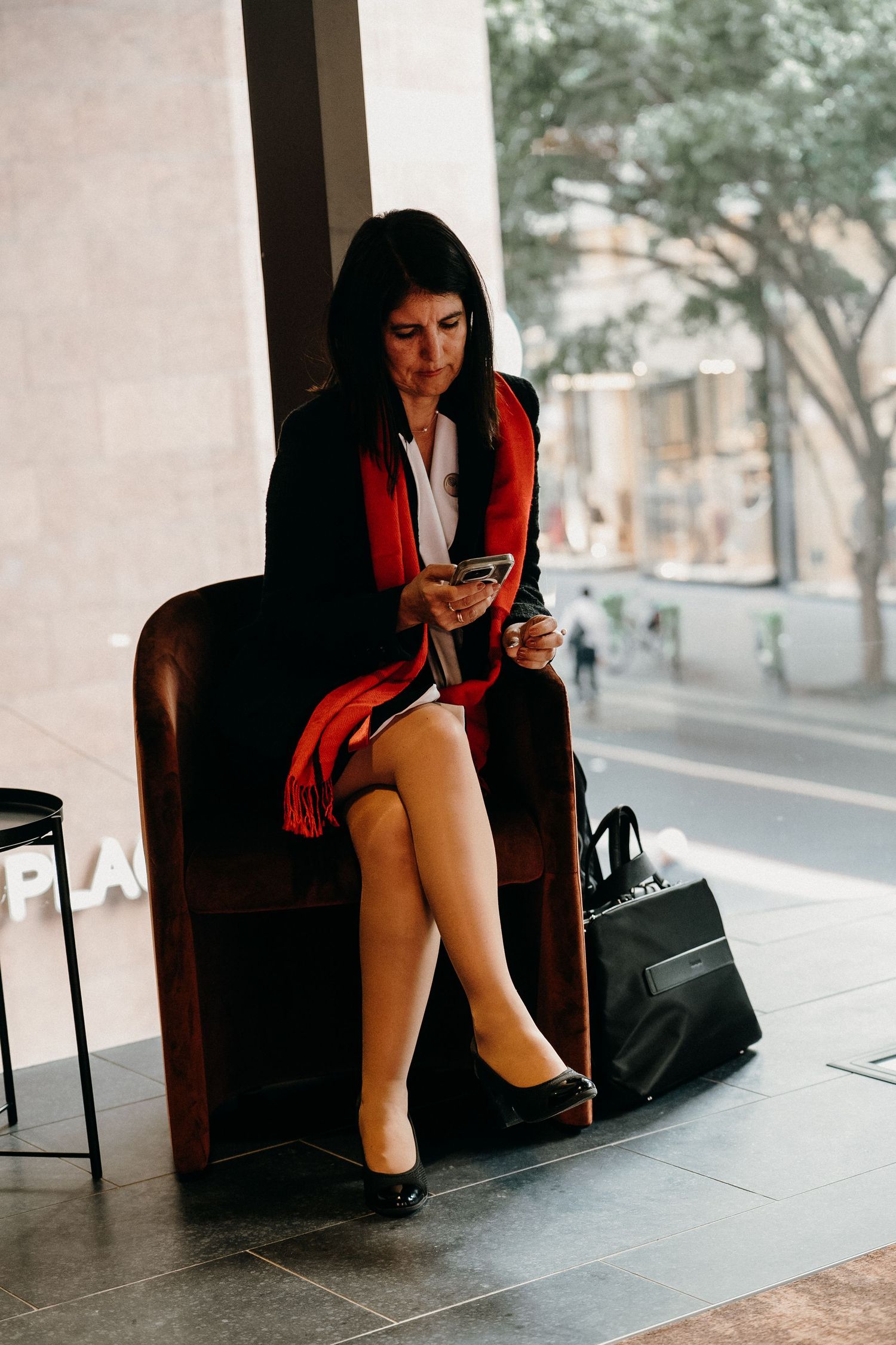 Professional in red blazer and skirt sitting on leather chair checking phone with handbag on floor.