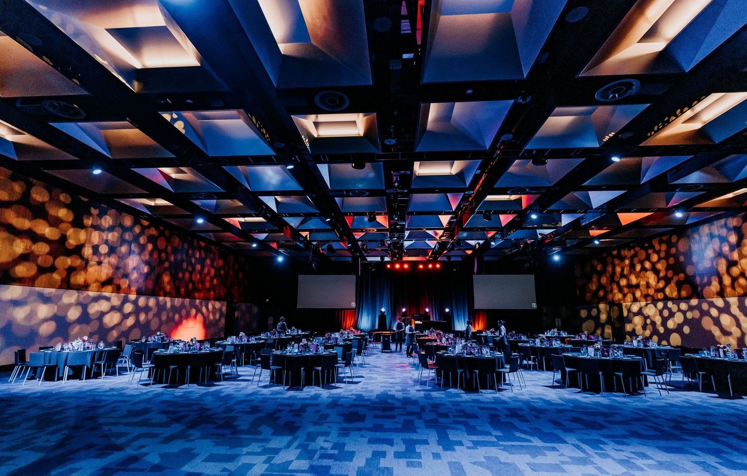 Large modern conference room with blue lighting and geometric ceiling design showcases rows of chairs for corporate events.