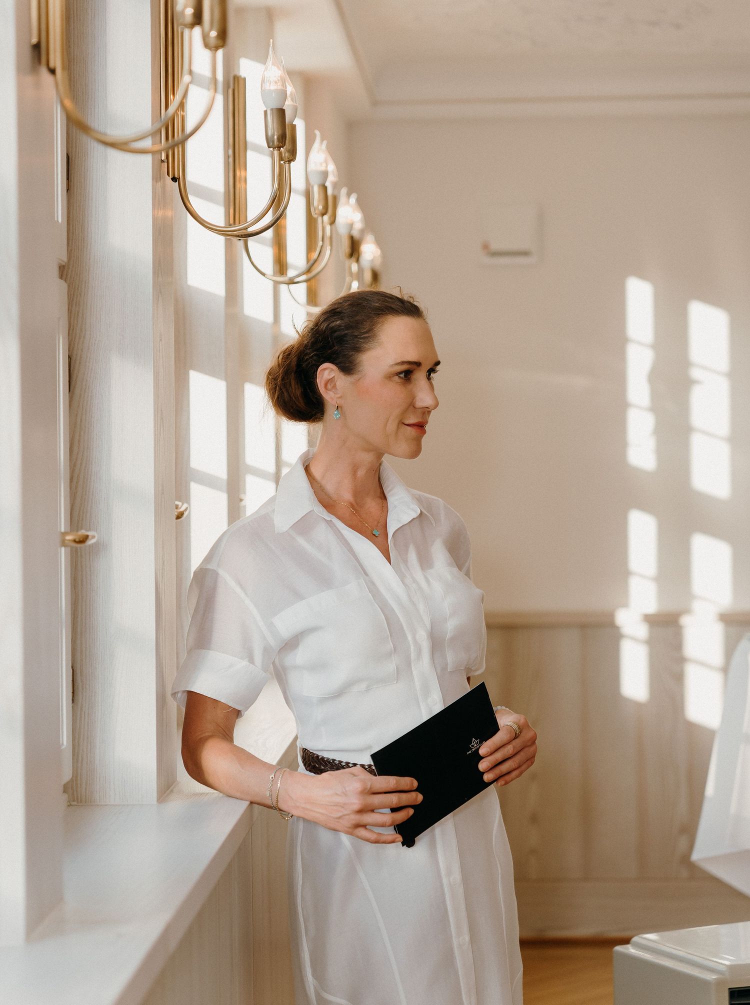 Woman in white dress holding a black notebook stands by sunlit windows with gold wall sconces.