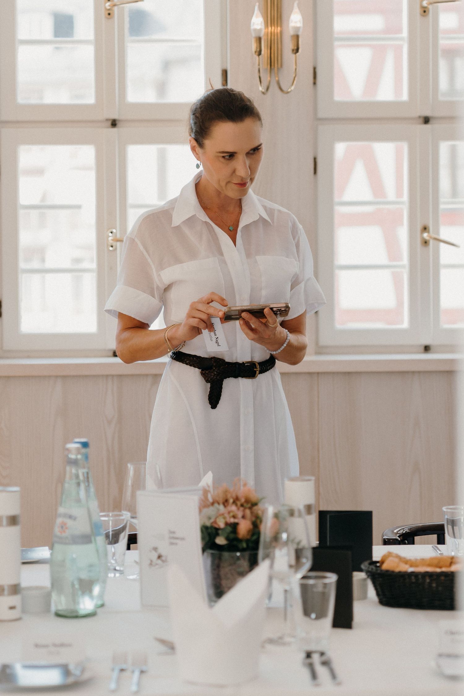 Woman in white dress using smartphone at elegantly set dining table in bright restaurant setting.