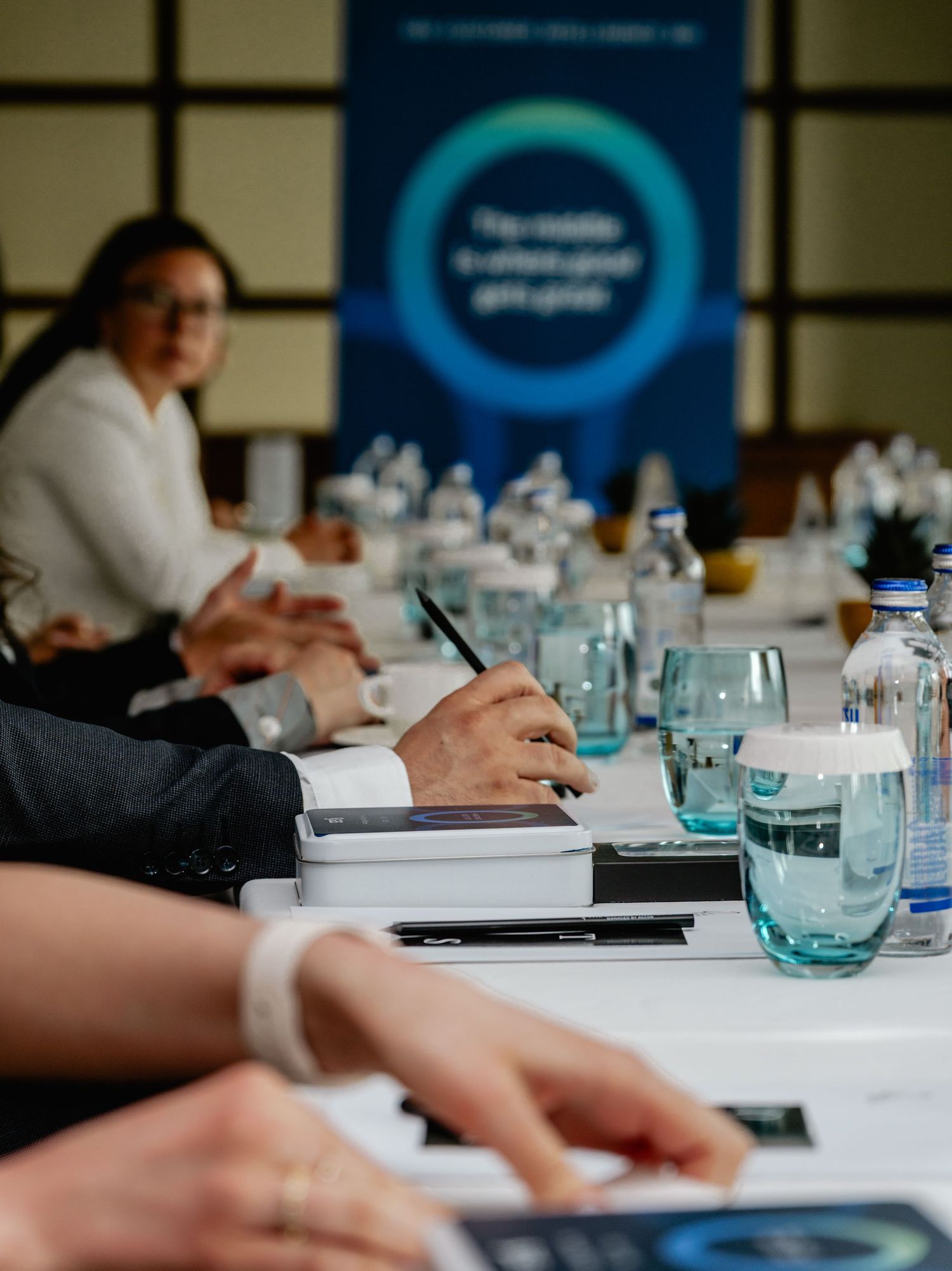 Business professionals at a conference table with water glasses, notebooks, and a blue branded banner in the background.