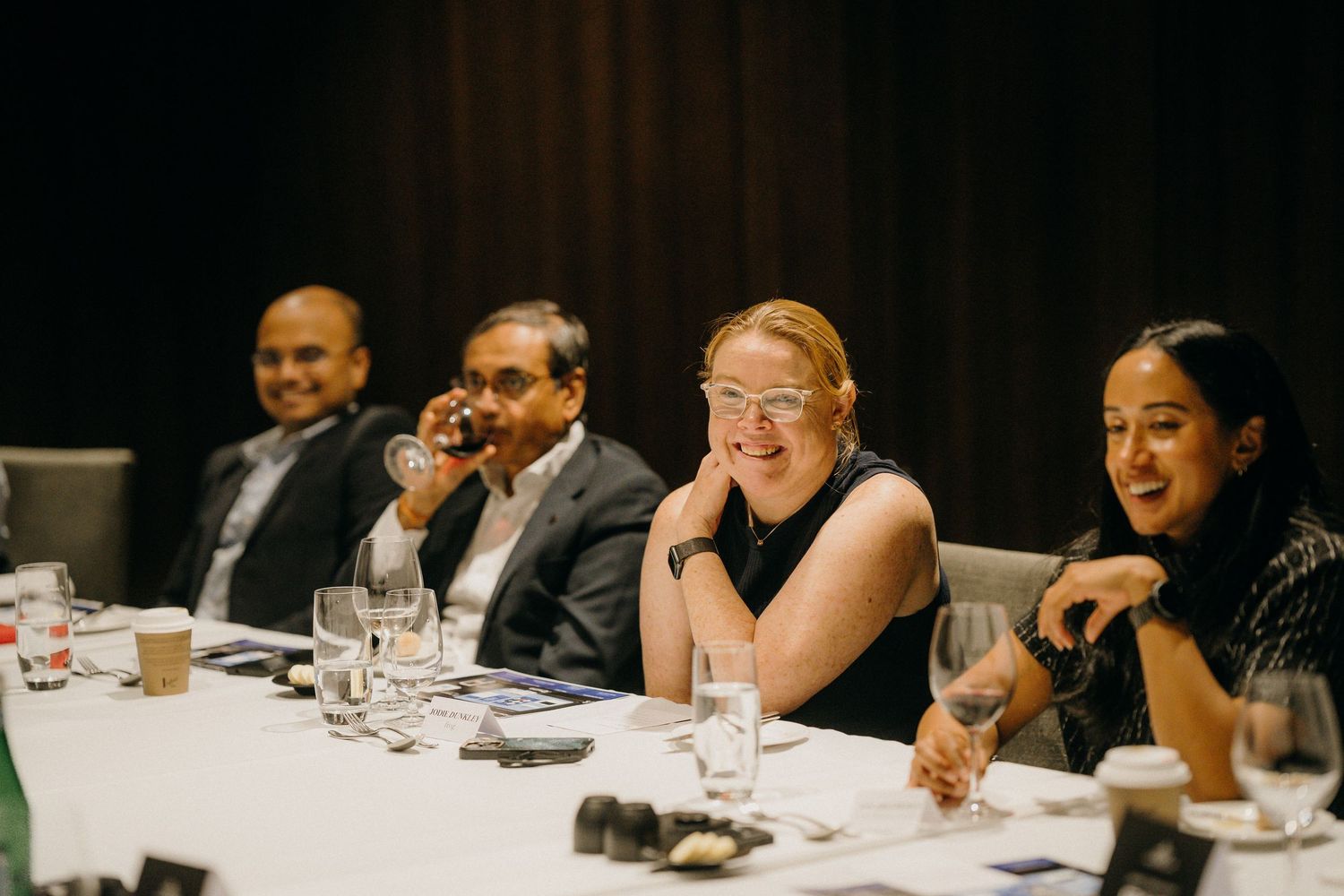 Four professionals laughing and socializing at a conference table during a formal business dinner event.