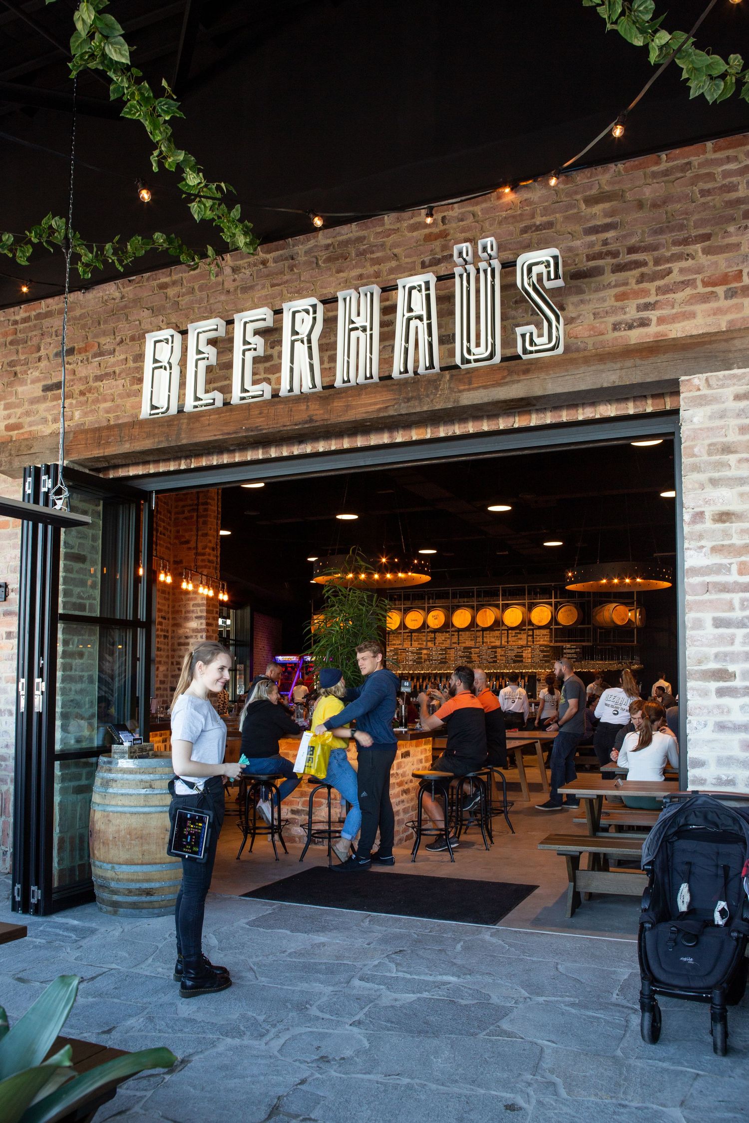 Modern beer hall with exposed brick walls and pendant lighting shows customers enjoying drinks at communal tables.