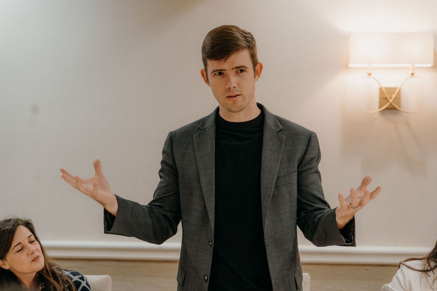 Professional in a gray suit and black shirt gestures while presenting in a modern office setting.