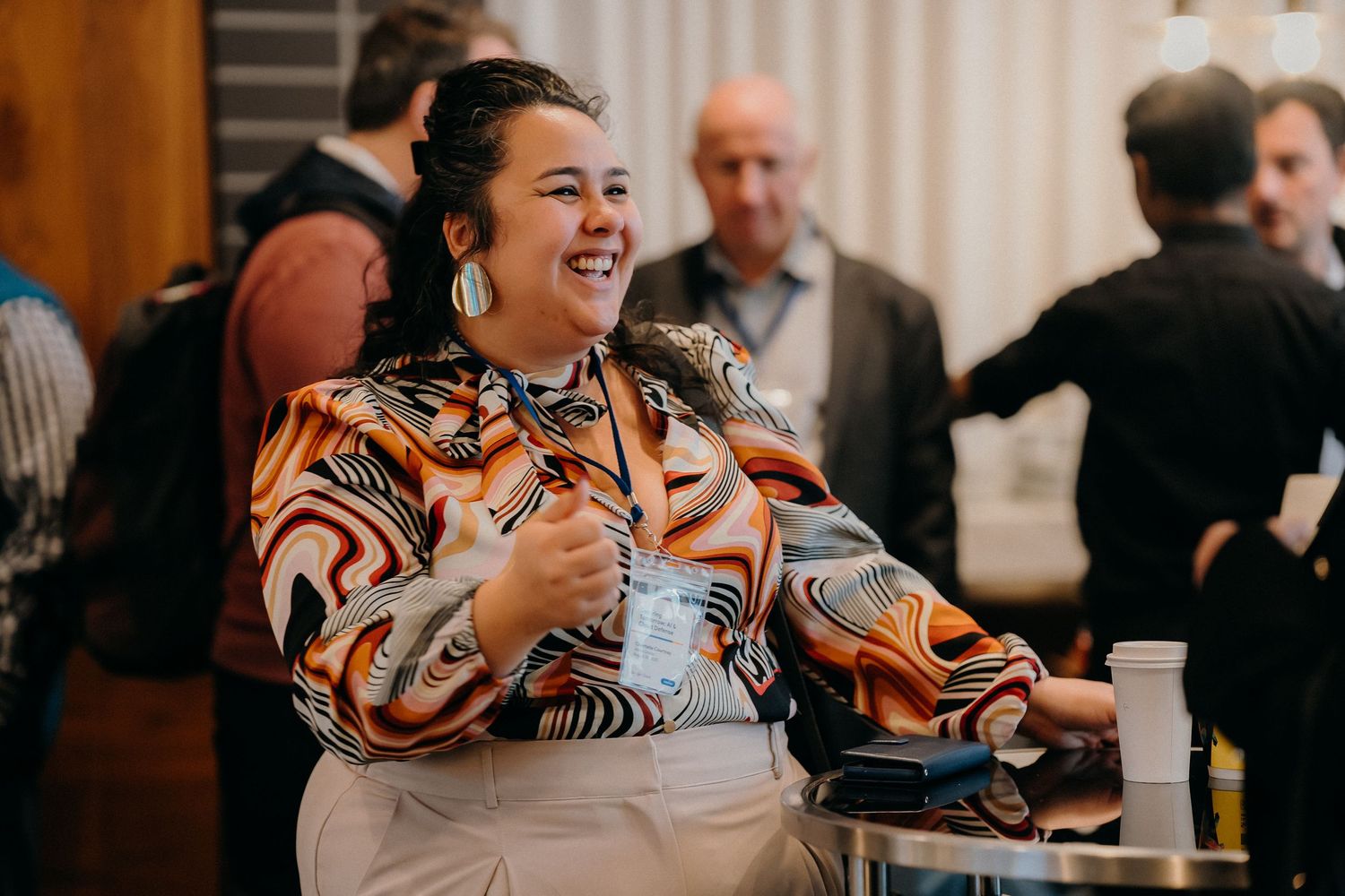 Someone wearing a colorful patterned shirt laughs joyfully while sitting in a room with other people in the background.