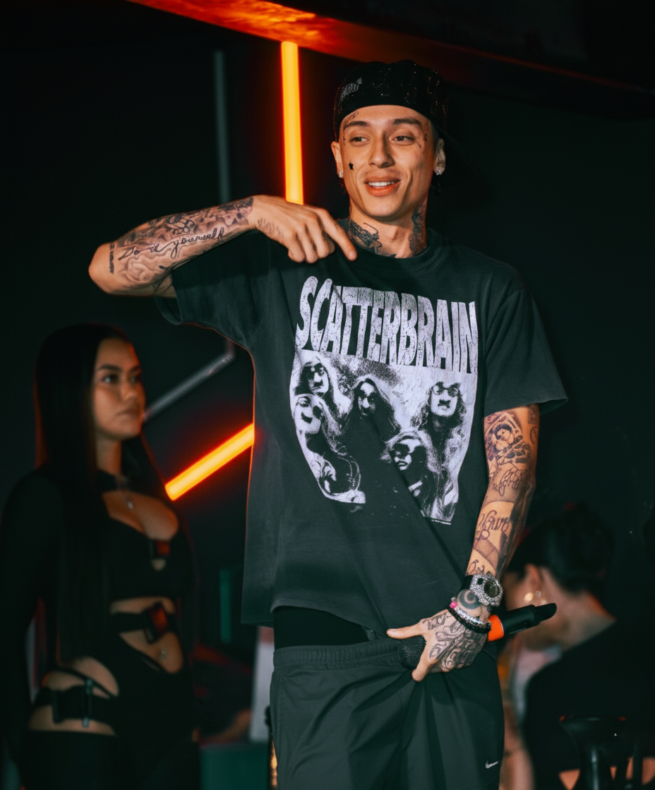 Person wearing a black Scatterbrain band t-shirt with tattoos on their arms performing on stage in dark lighting.