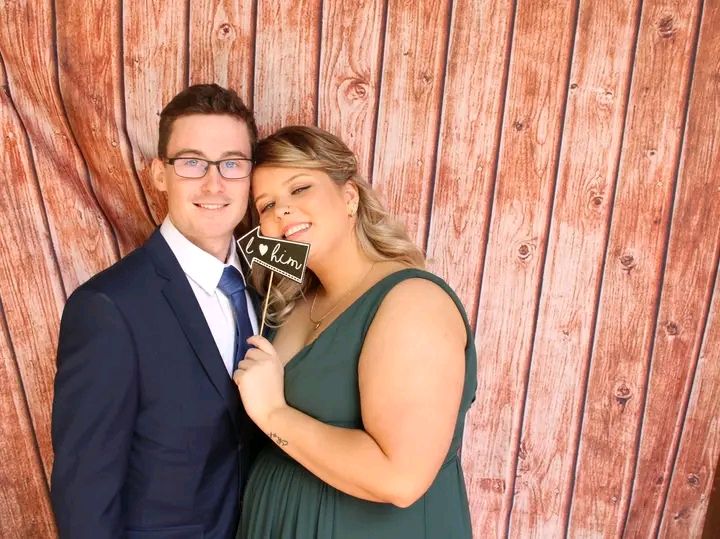 A couple poses together against a rustic wooden wall background, with one wearing a navy suit and one in a green dress.