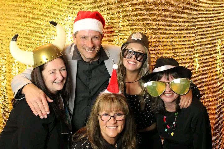Group of people wearing festive holiday attire smile and pose together against a shimmering gold backdrop.