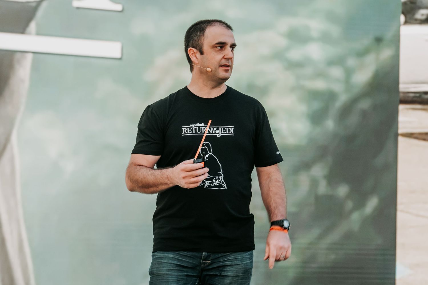 Person in black t-shirt holding phone while standing against light colored wall with blurred background.