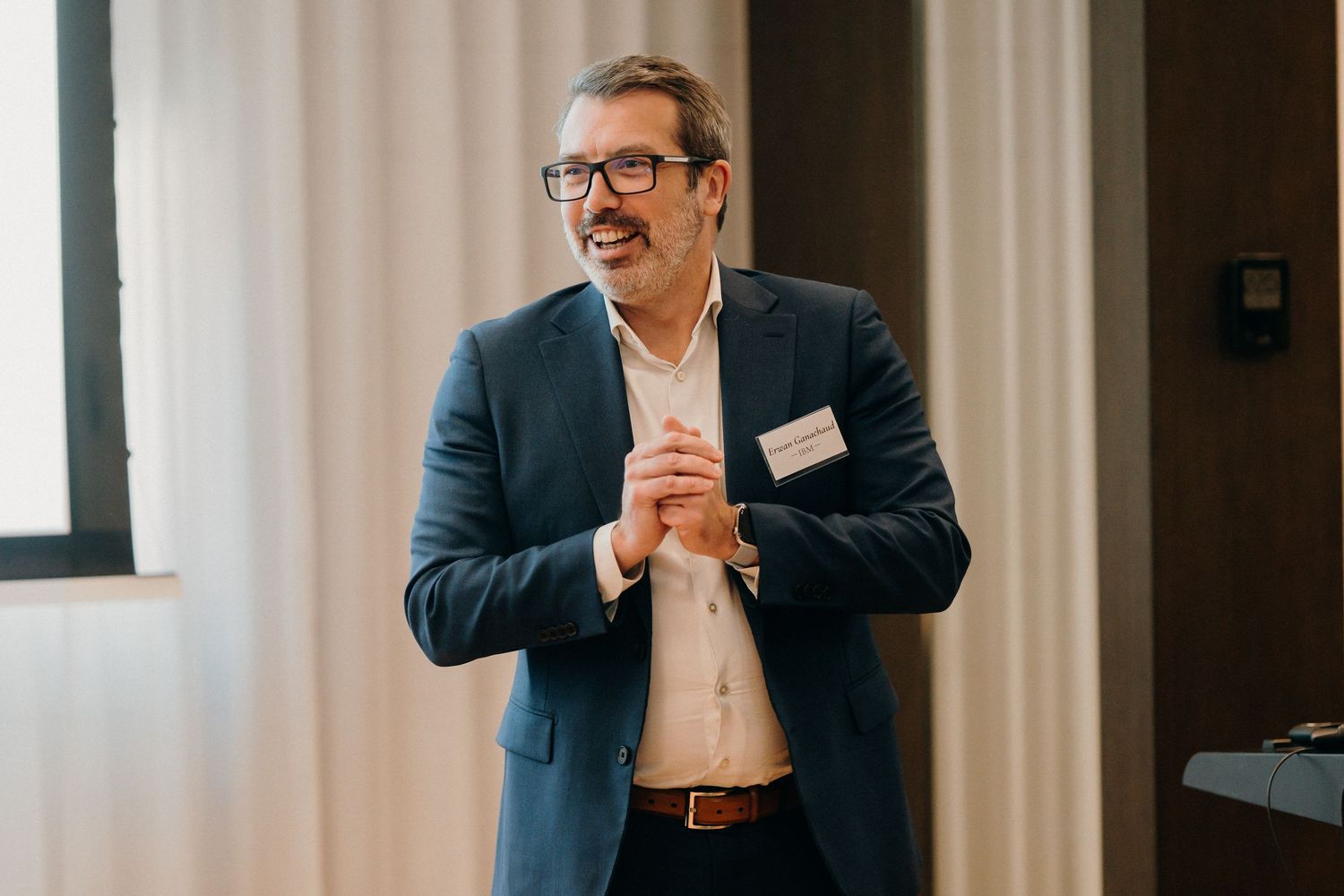 Professional in a dark suit and glasses gestures while speaking during a business presentation.