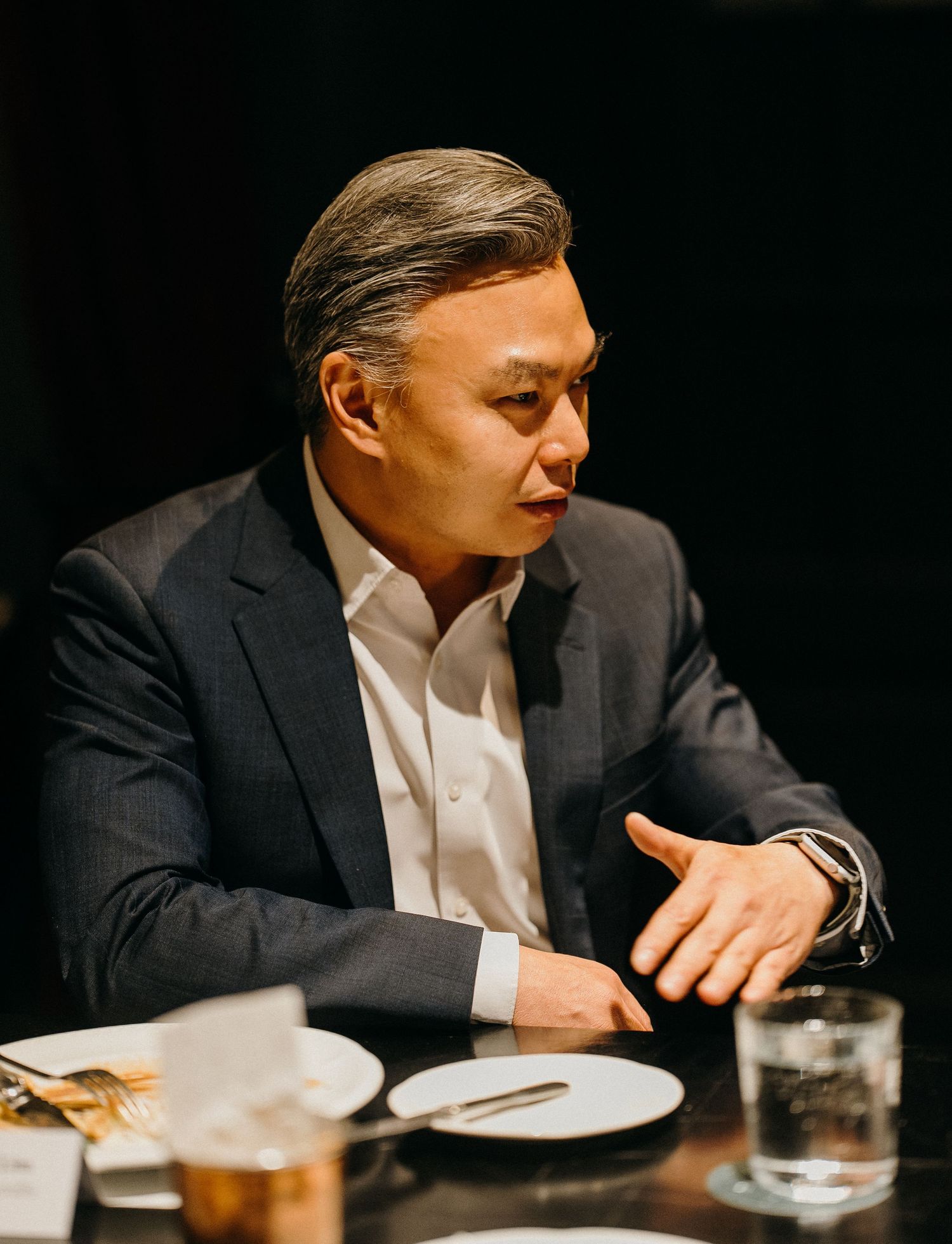A person in a dark suit gestures while sitting at a dining table with plates and drinks.