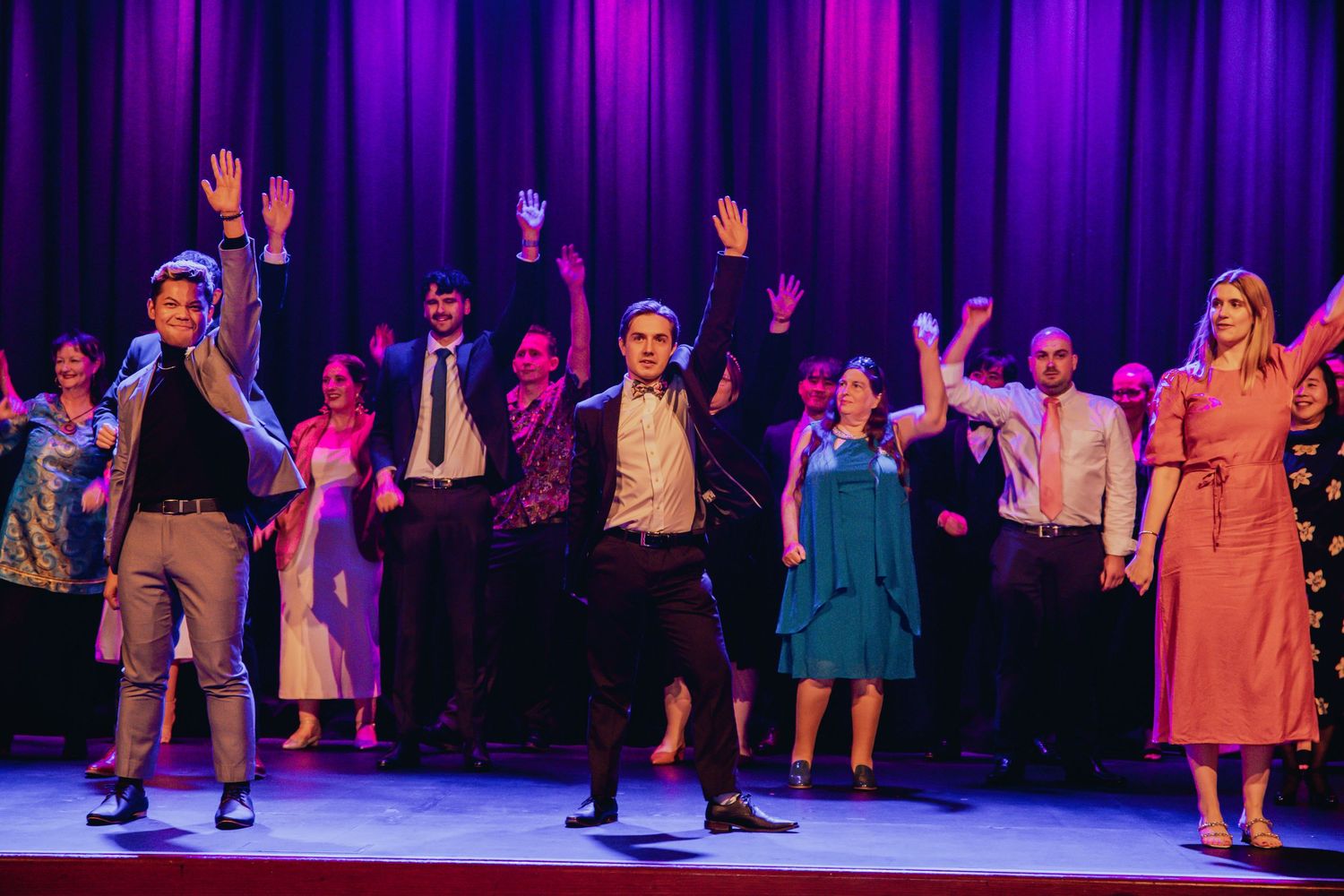 Theater performers on stage with raised arms during a musical performance with a purple curtain backdrop.