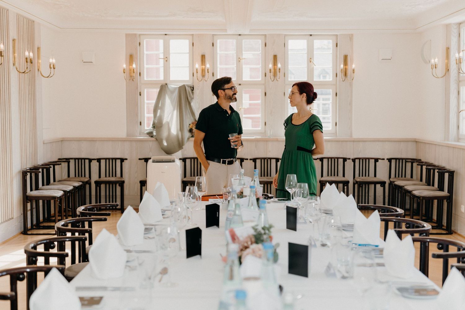 Elegant wedding reception hall with long white tables set for dining, two guests conversing near large windows.