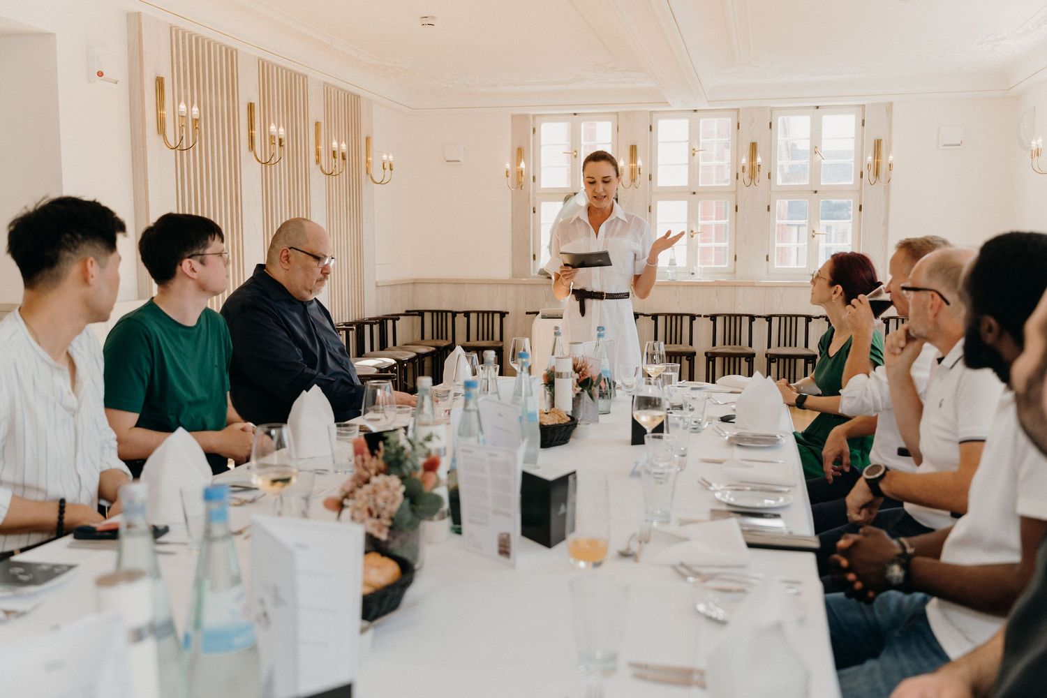 A diverse group of people seated at a long dining table listen to a presenter speaking at an elegant restaurant.