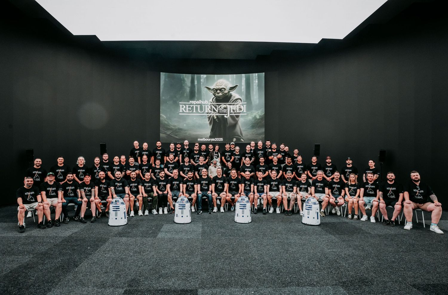 Large group photo of team members in black uniforms seated together in front of a dark wall with a projected logo.