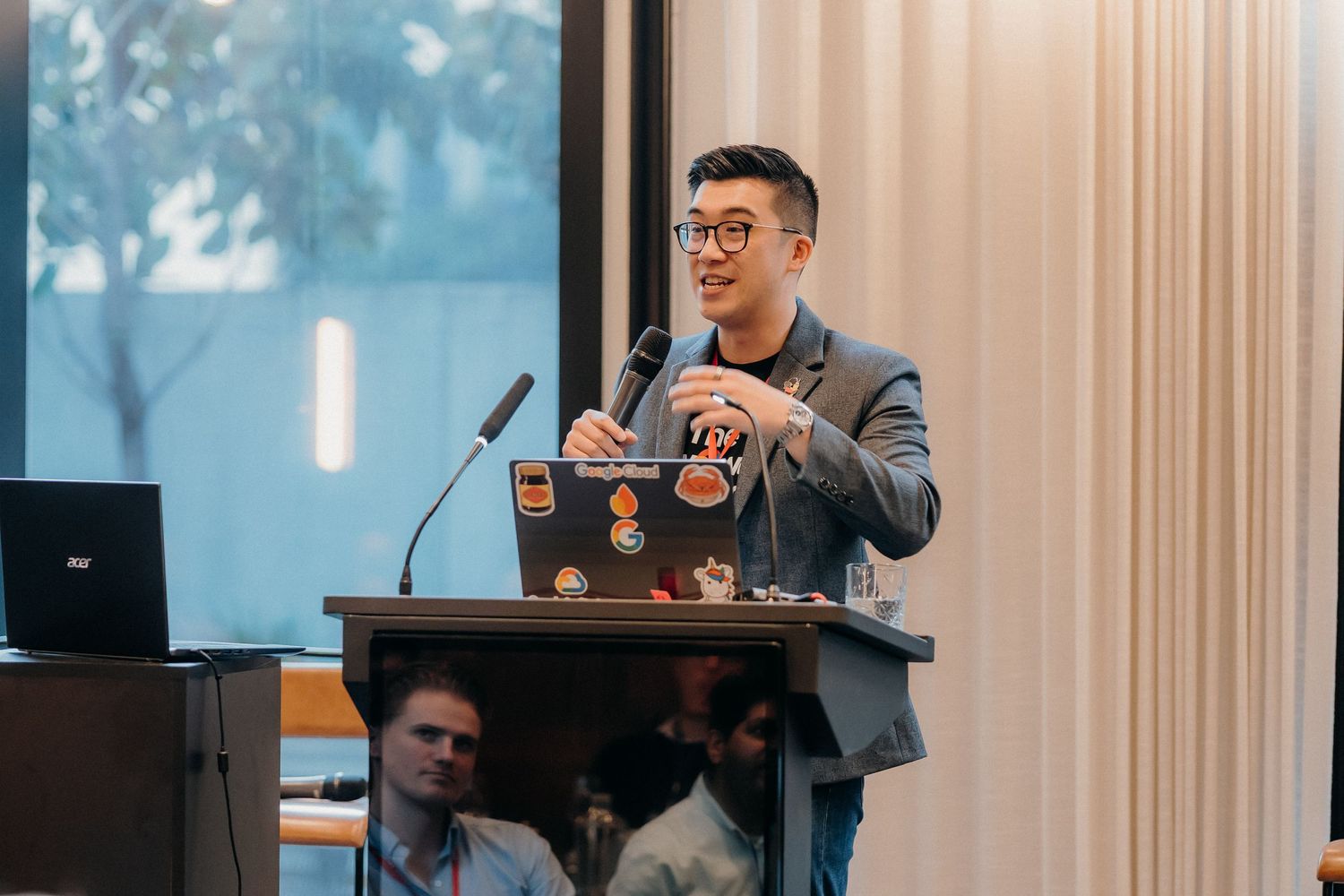A speaker gestures enthusiastically while presenting at a business conference podium with curtains in the background.