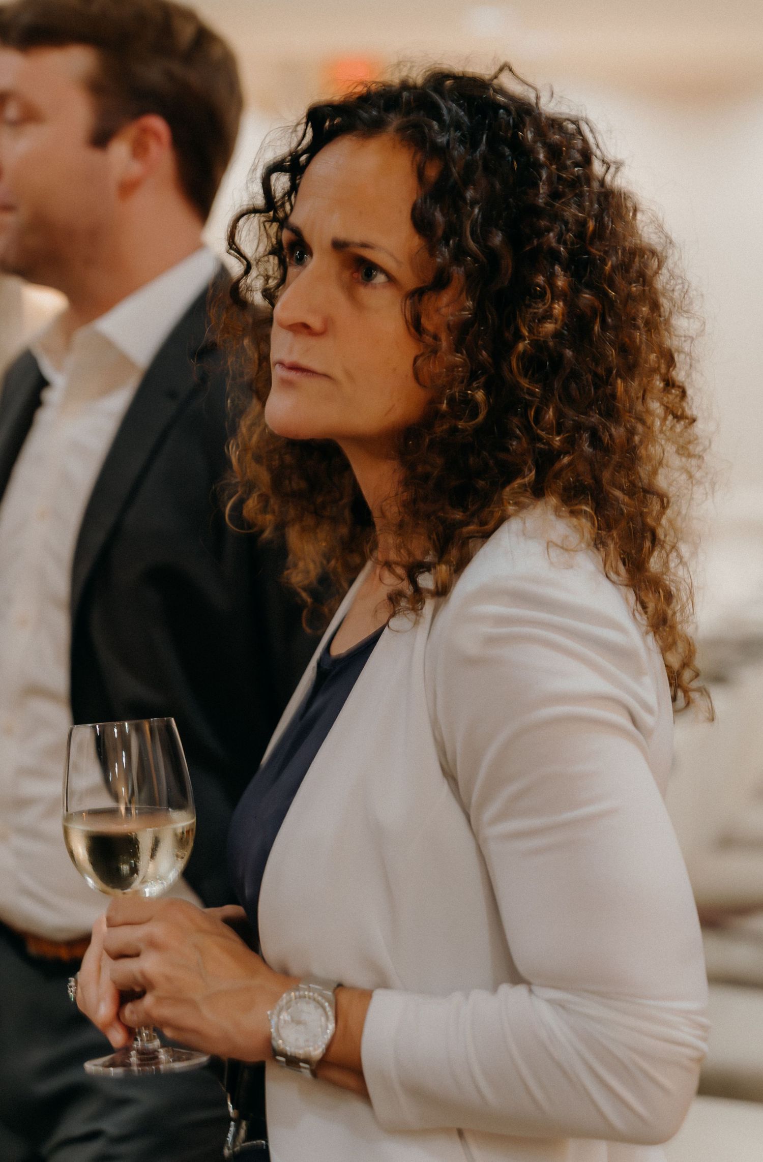 Person in white blazer holding wine glass at formal event with curly hair and elegant attire.