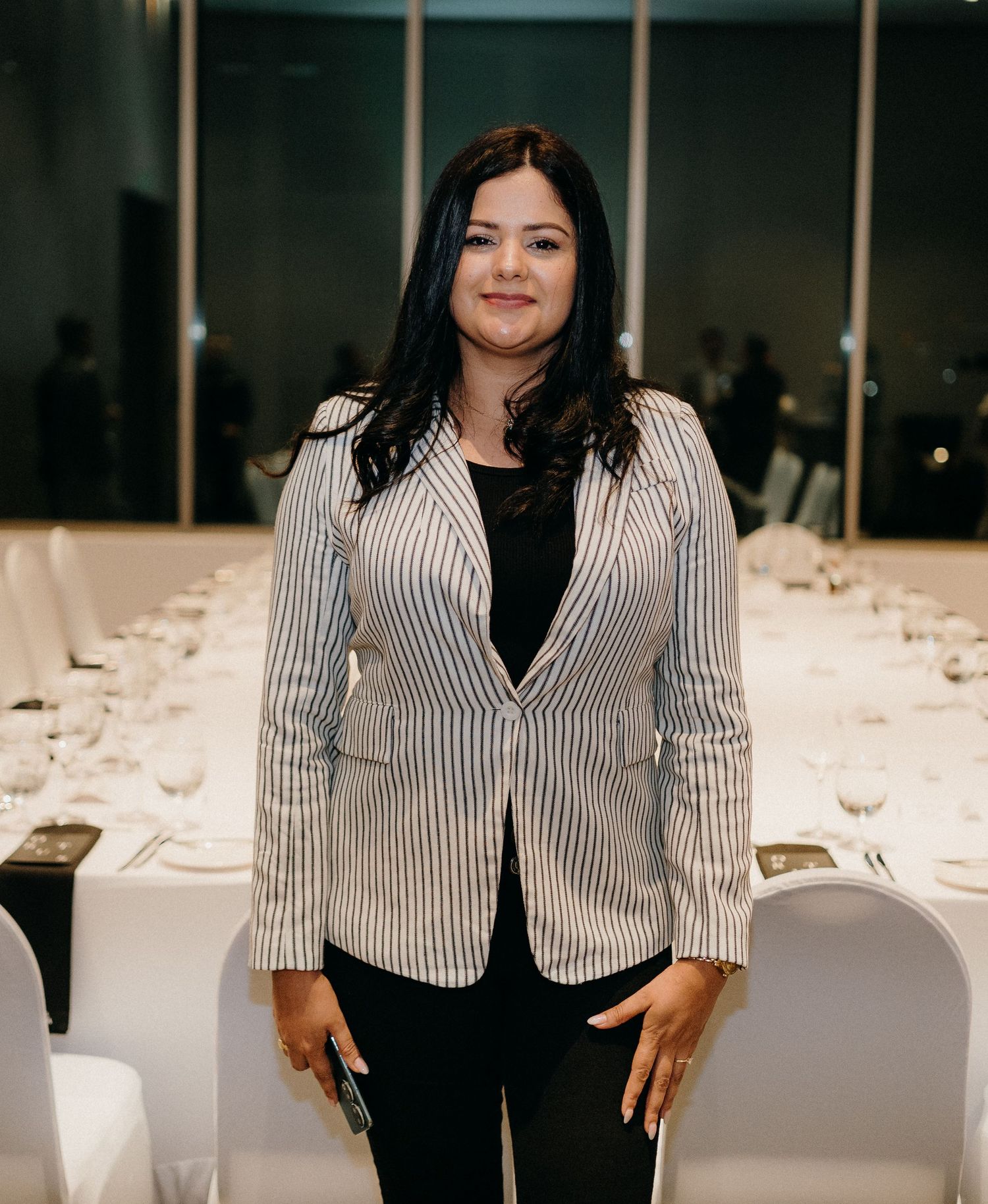 A person in a striped blazer stands in front of a formal dining setup with white tablecloths and place settings.