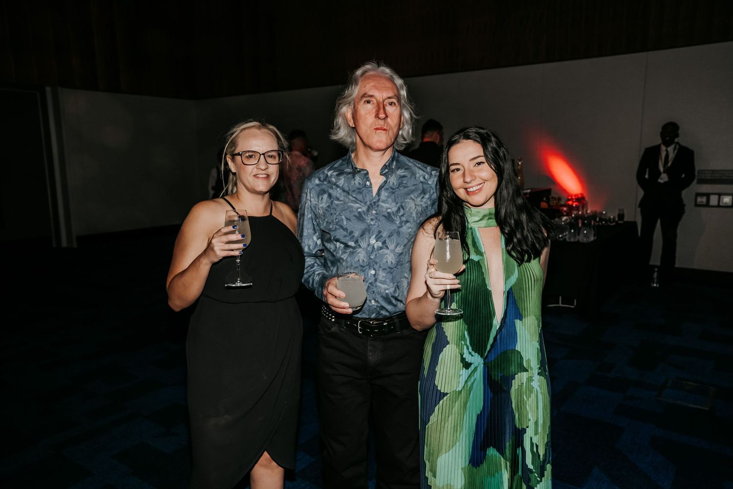 Three people pose together at a dark event space, with one wearing a blue shirt and another in a green dress.