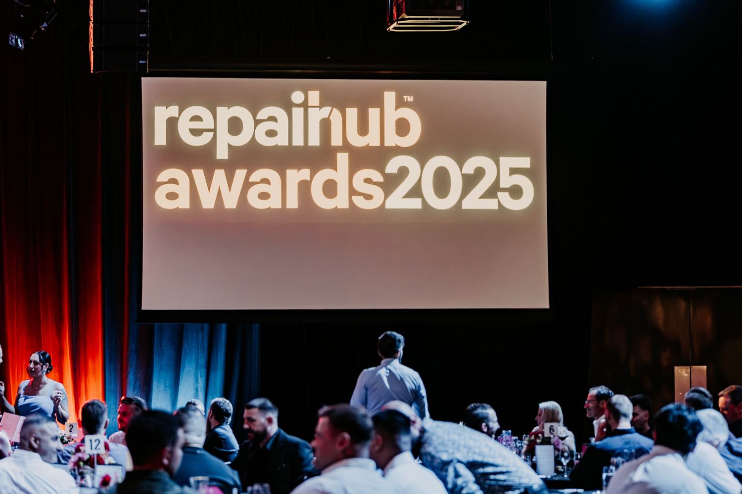 Conference attendees gather in an auditorium with red and blue lighting while viewing a presentation on screen.