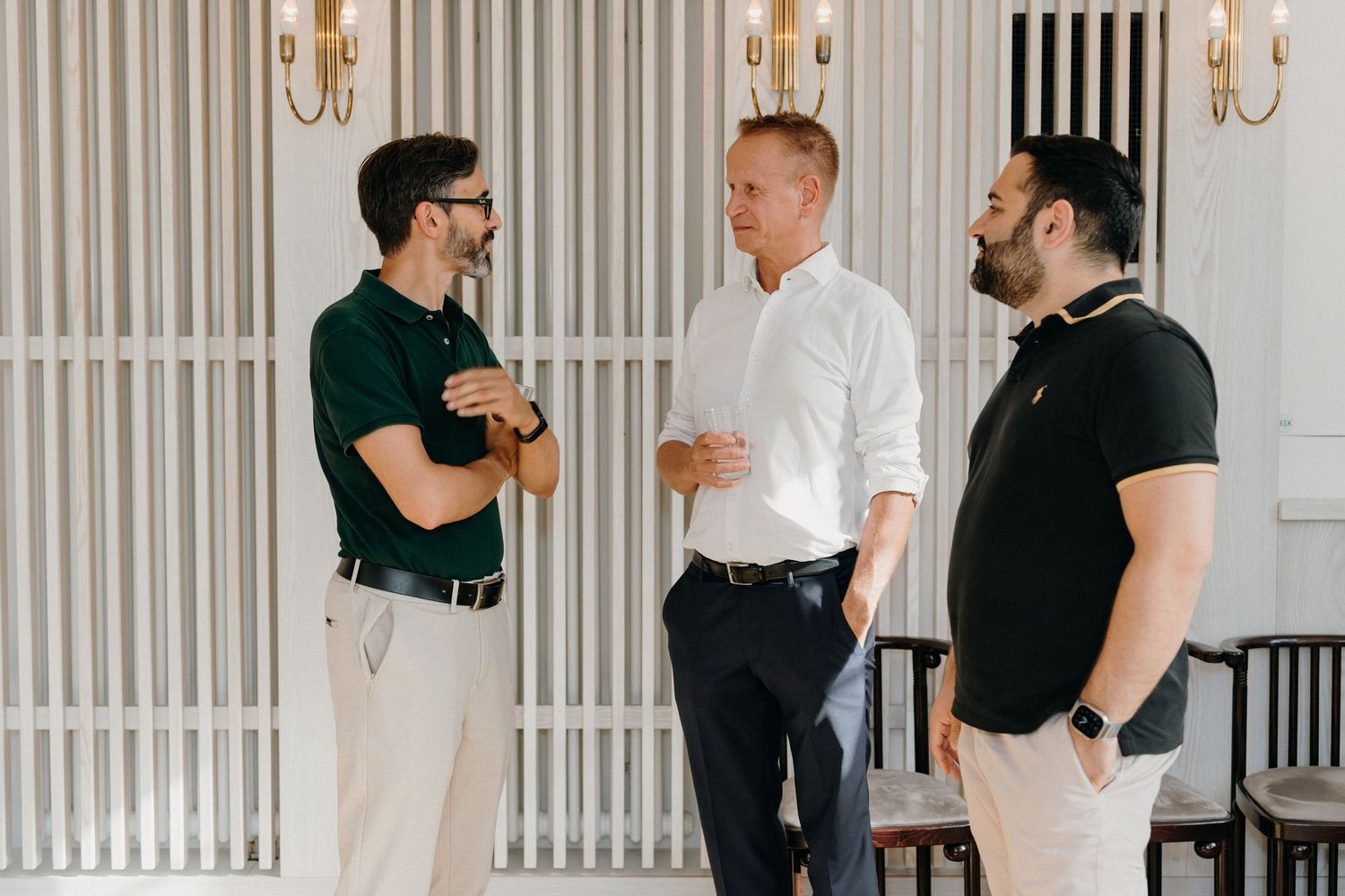 Three men in smart casual attire engaged in conversation indoors near elegant white paneled walls with wall sconces.
