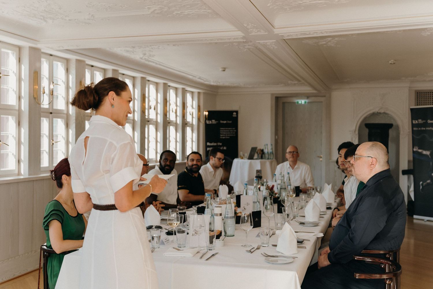 Bride in white dress giving speech at elegant wedding reception dinner table with seated guests.