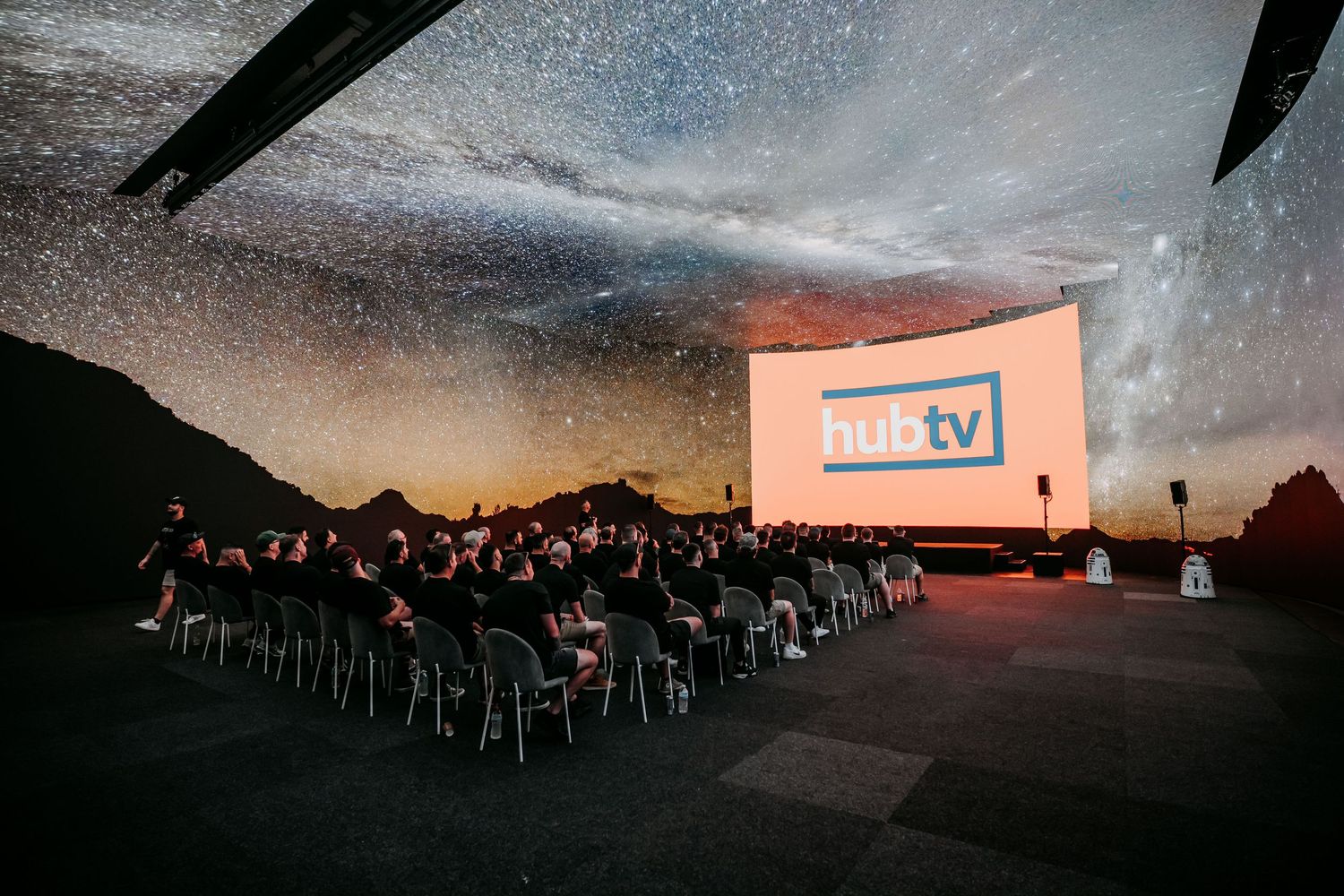 A crowd watches a movie at an outdoor cinema under a starry night sky with pink sunset glow.