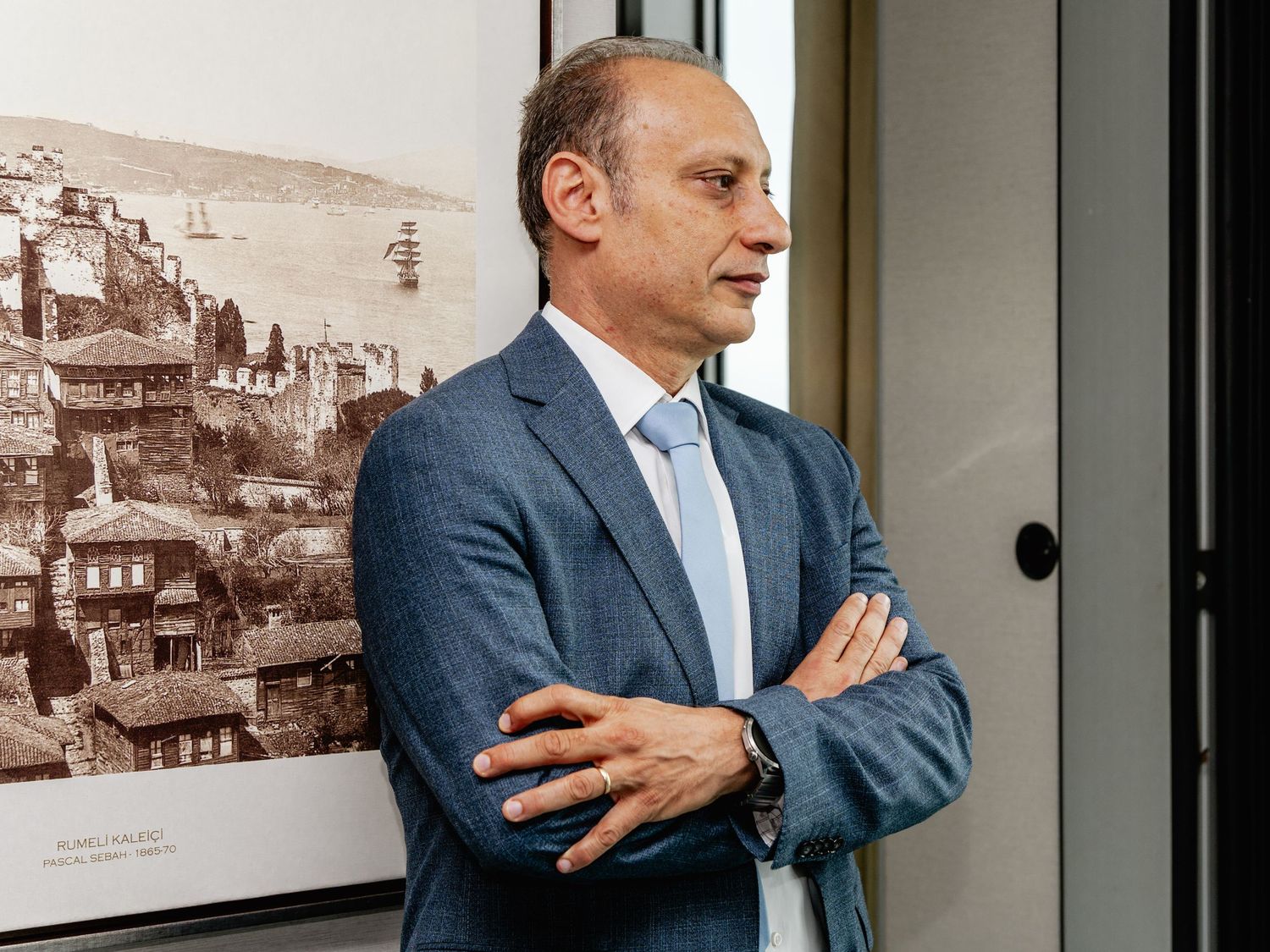 Professional man in blue suit with arms crossed standing near vintage cityscape photograph on wall.