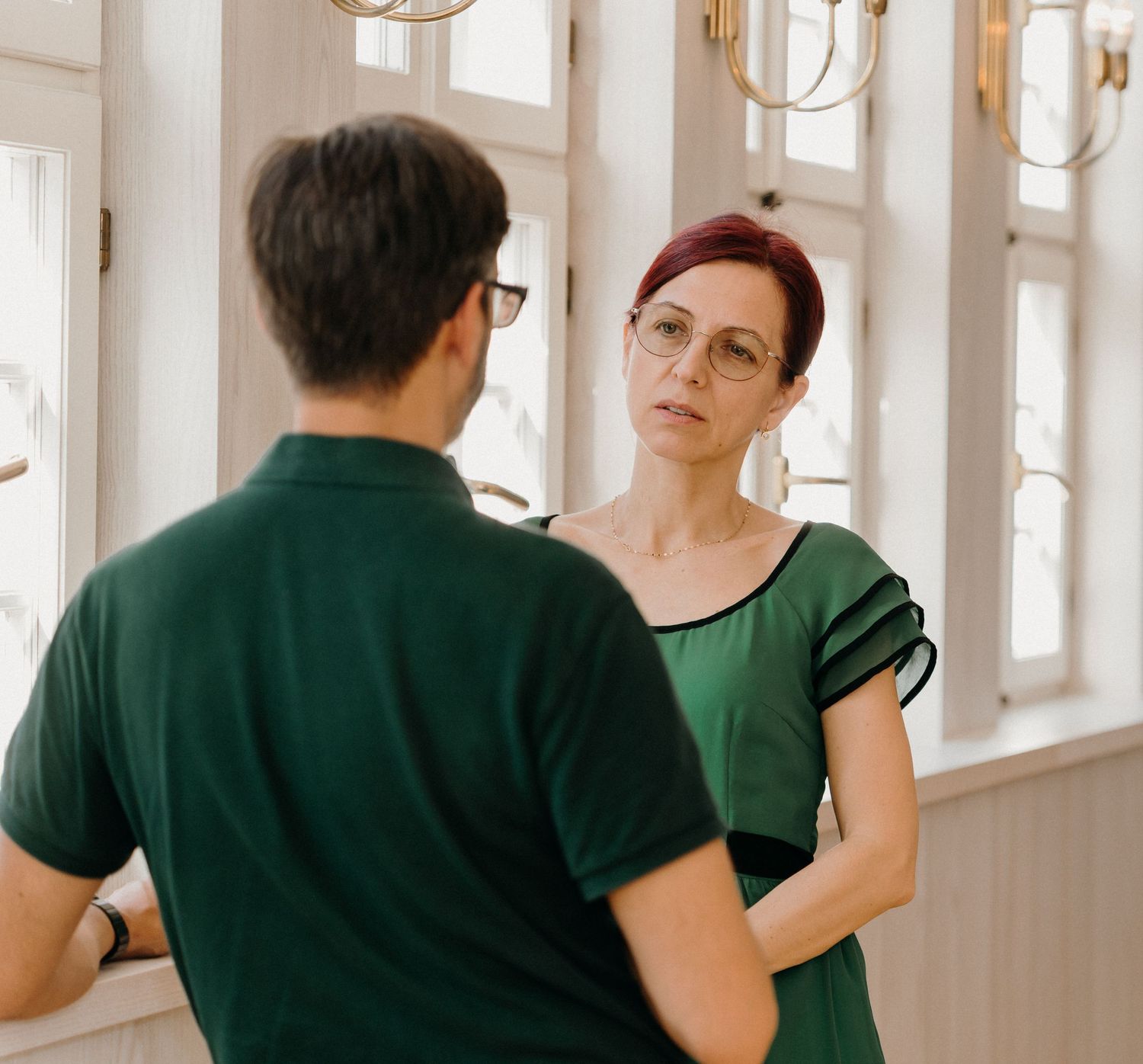 Two people wearing green outfits having a conversation indoors near bright windows in an elegant room.