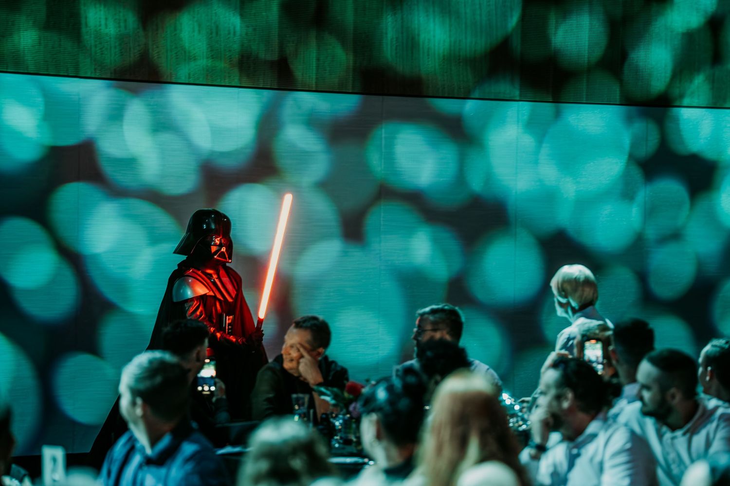 A Star Wars cosplayer dressed as Darth Vader holds a lightsaber amidst a crowd with turquoise bokeh lighting effects.