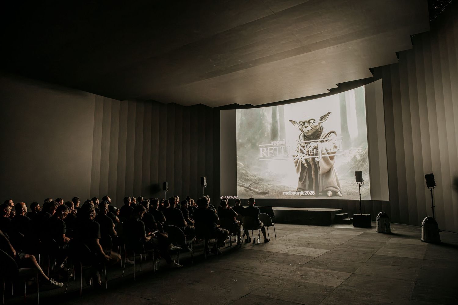 A cinematic screening room with rows of seats and a large white projection screen showing a silhouetted figure.