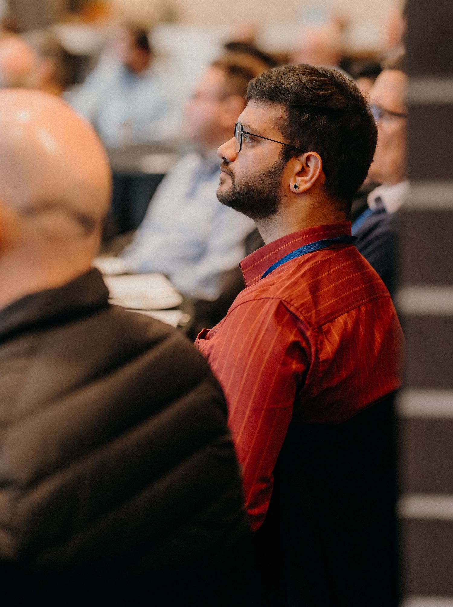 A person wearing glasses and a red striped shirt sits attentively in an audience during a presentation.
