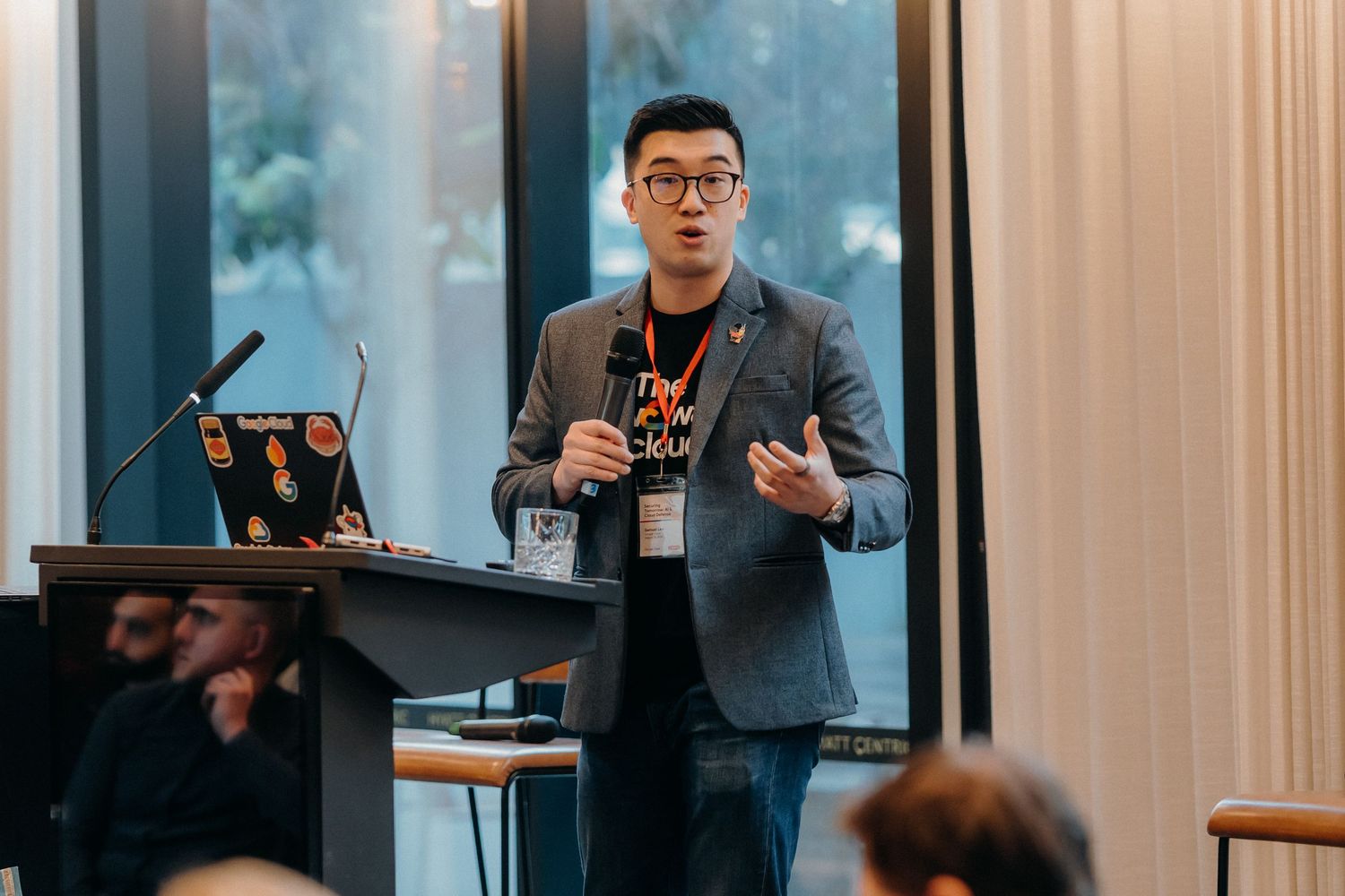A speaker in a gray blazer presents at a podium during a professional conference or seminar.