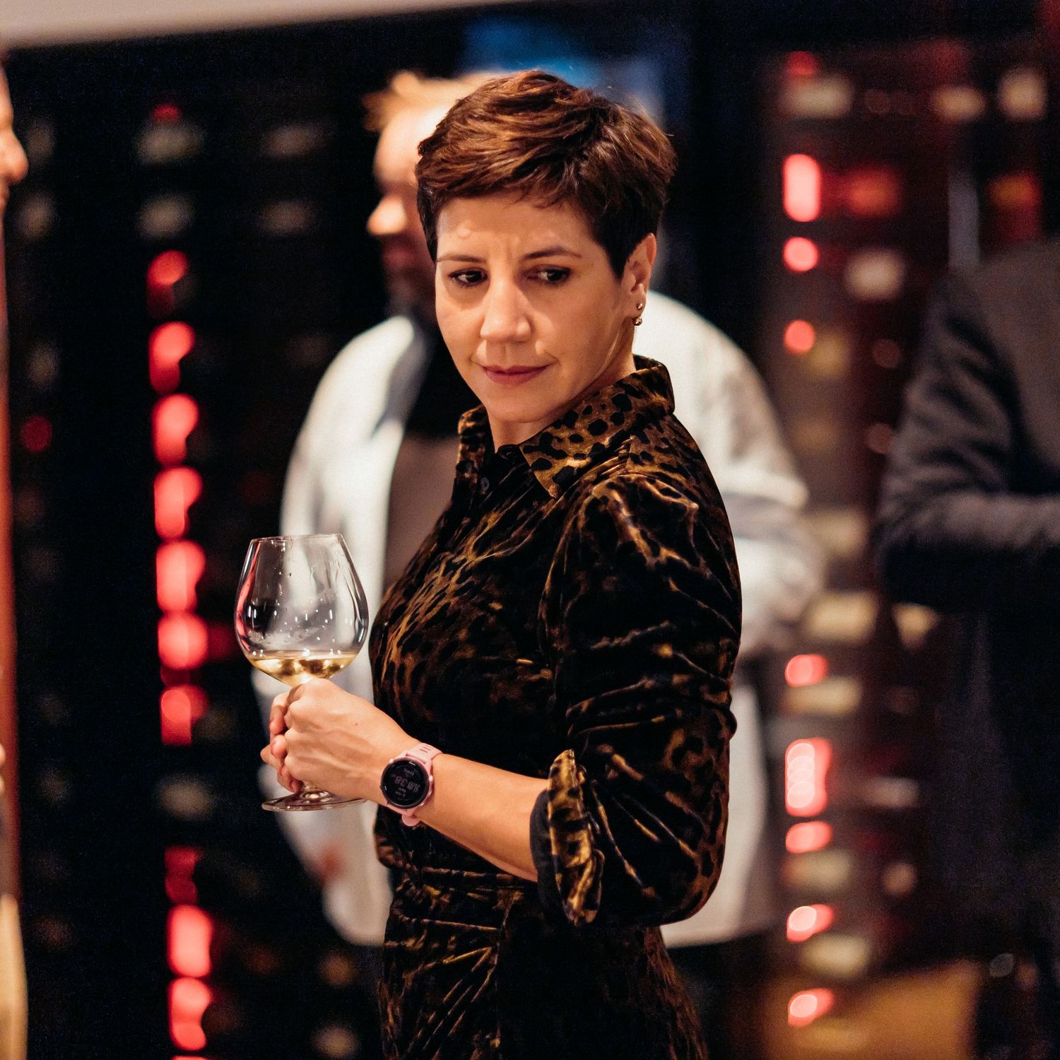 Person in black attire holds wine glass while seated at dimly lit venue with red ambient lighting in background.