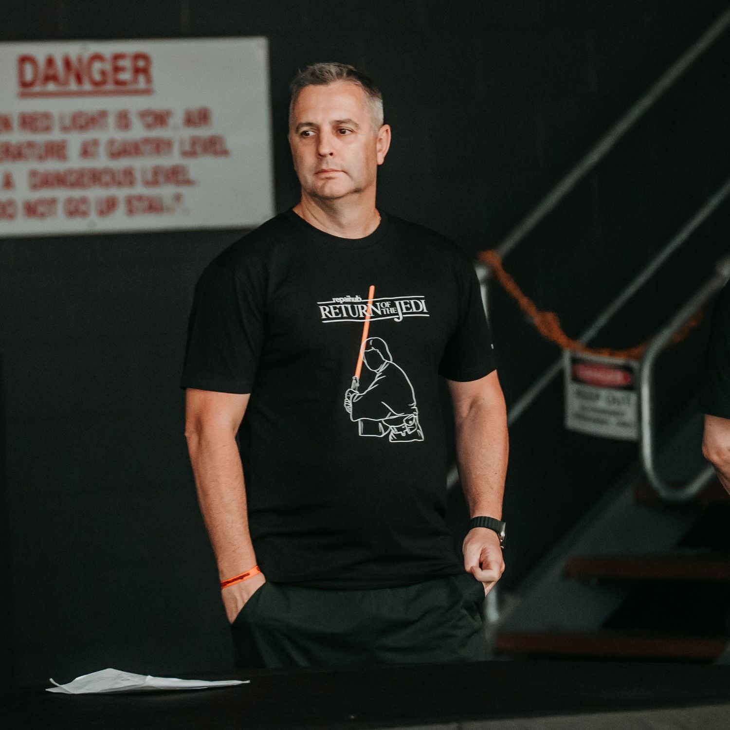 A person in a black t-shirt sits in a dark room with danger signs visible on the wall behind them.