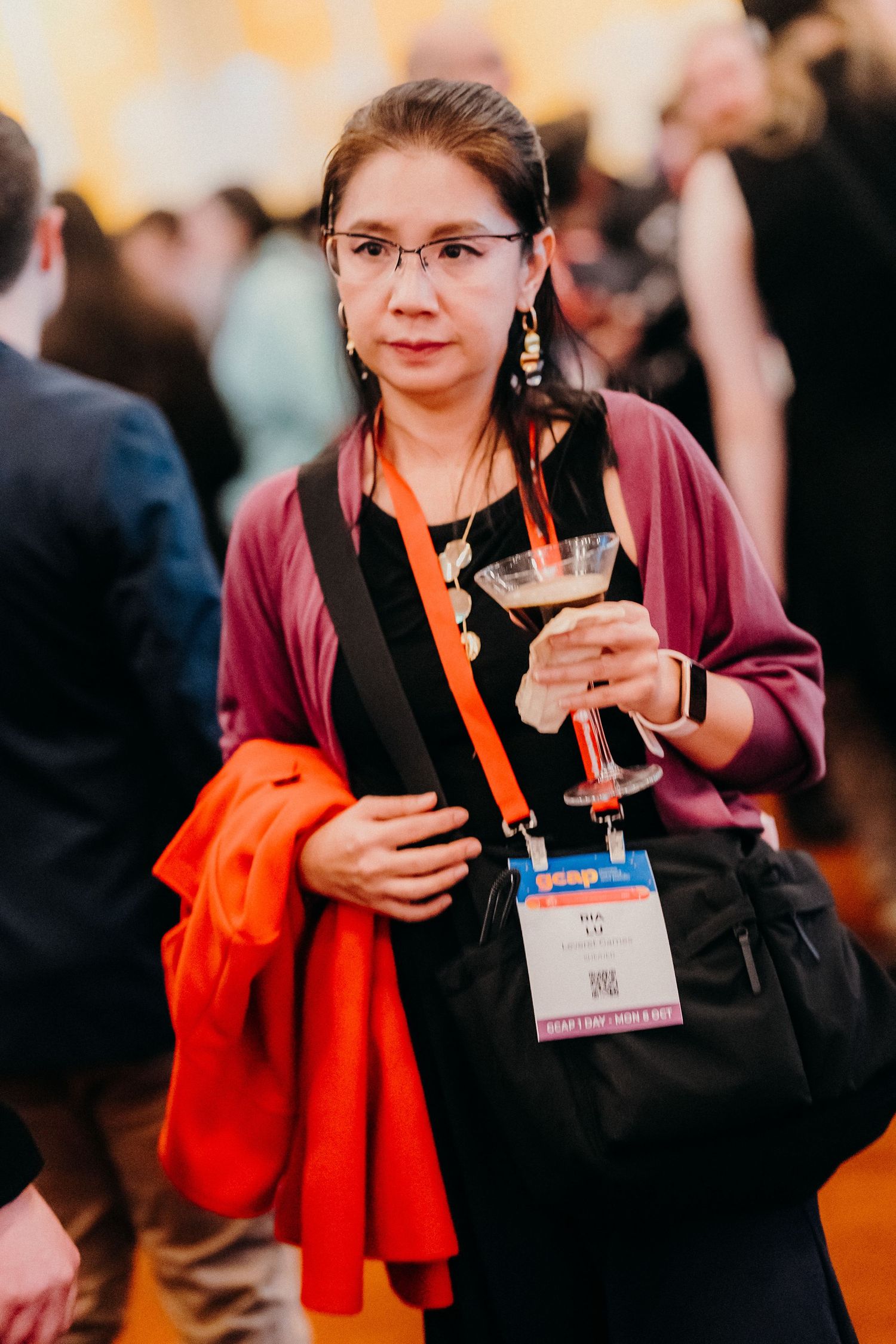 A person in a burgundy sweater and orange scarf smiles while holding a conference badge and beverage at an event.