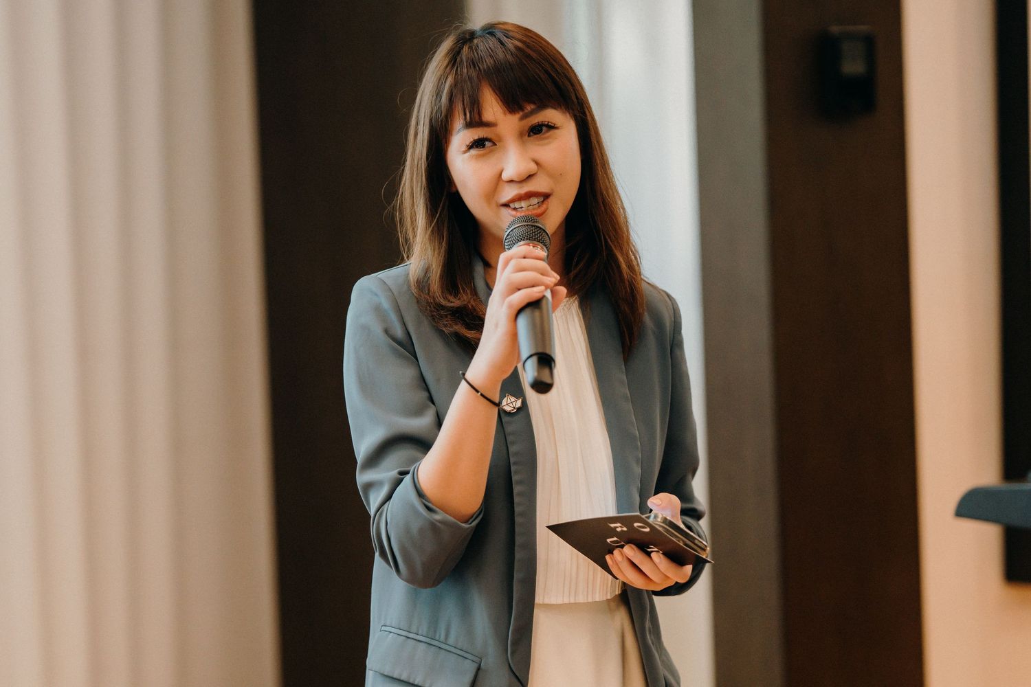 A person in a gray blazer speaks into a microphone while gesturing during a presentation.
