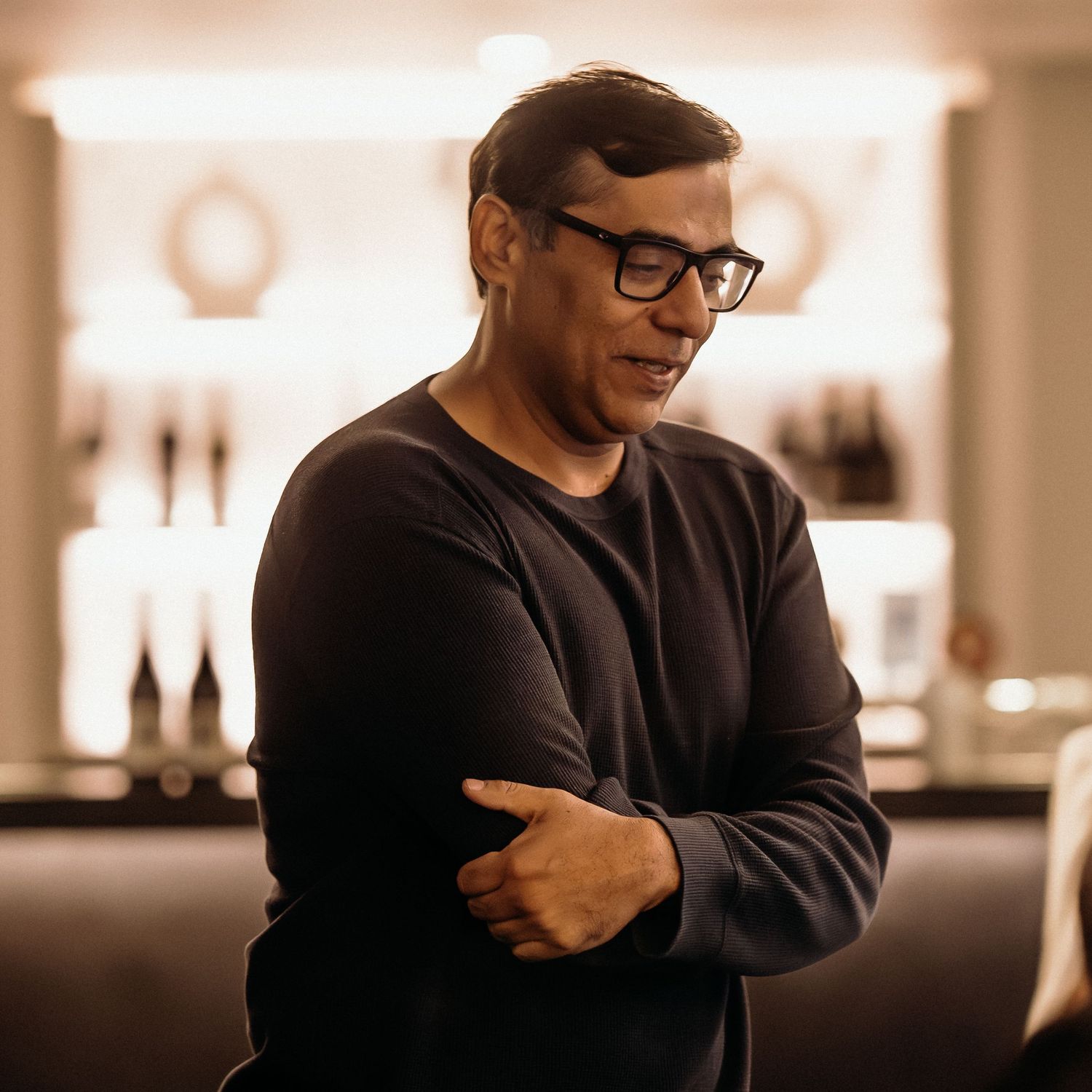 Person wearing glasses and a black sweater stands with arms crossed in warm-lit indoor setting.