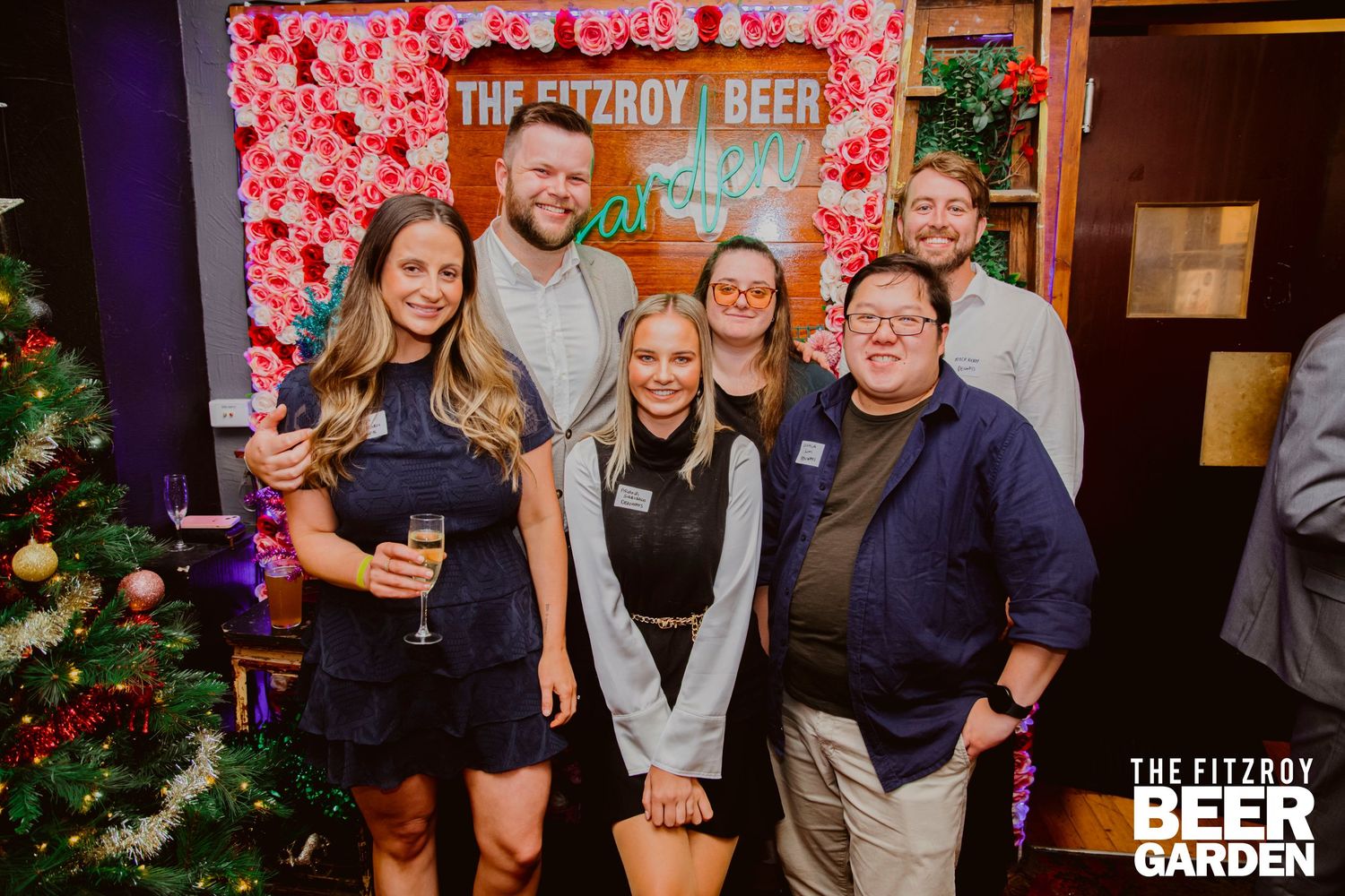 A group of people gathered at a festive holiday party with red decorative lighting and Christmas trees in the background.
