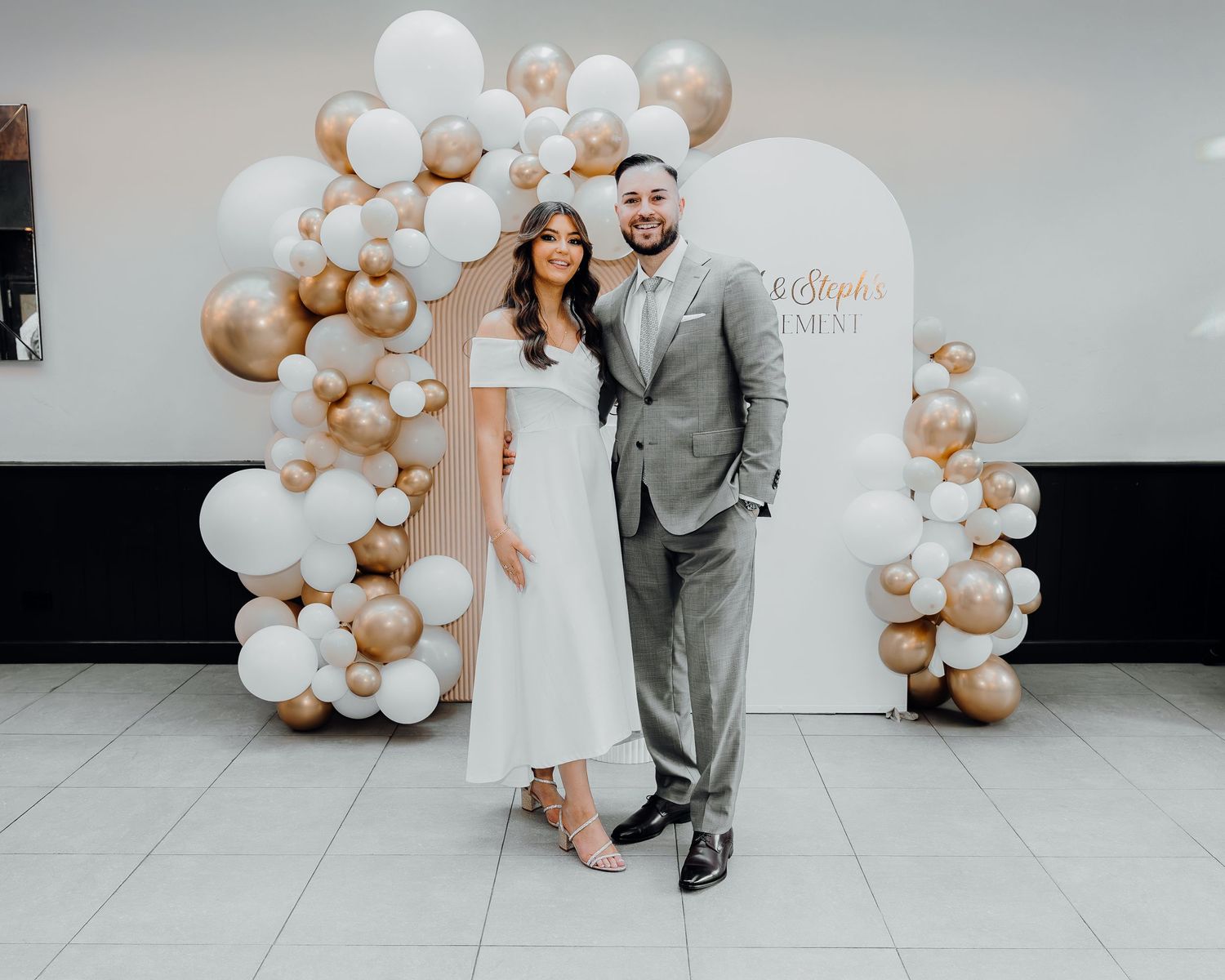 A couple poses in front of an elegant balloon arch decorated in white and gold colors at their wedding celebration.