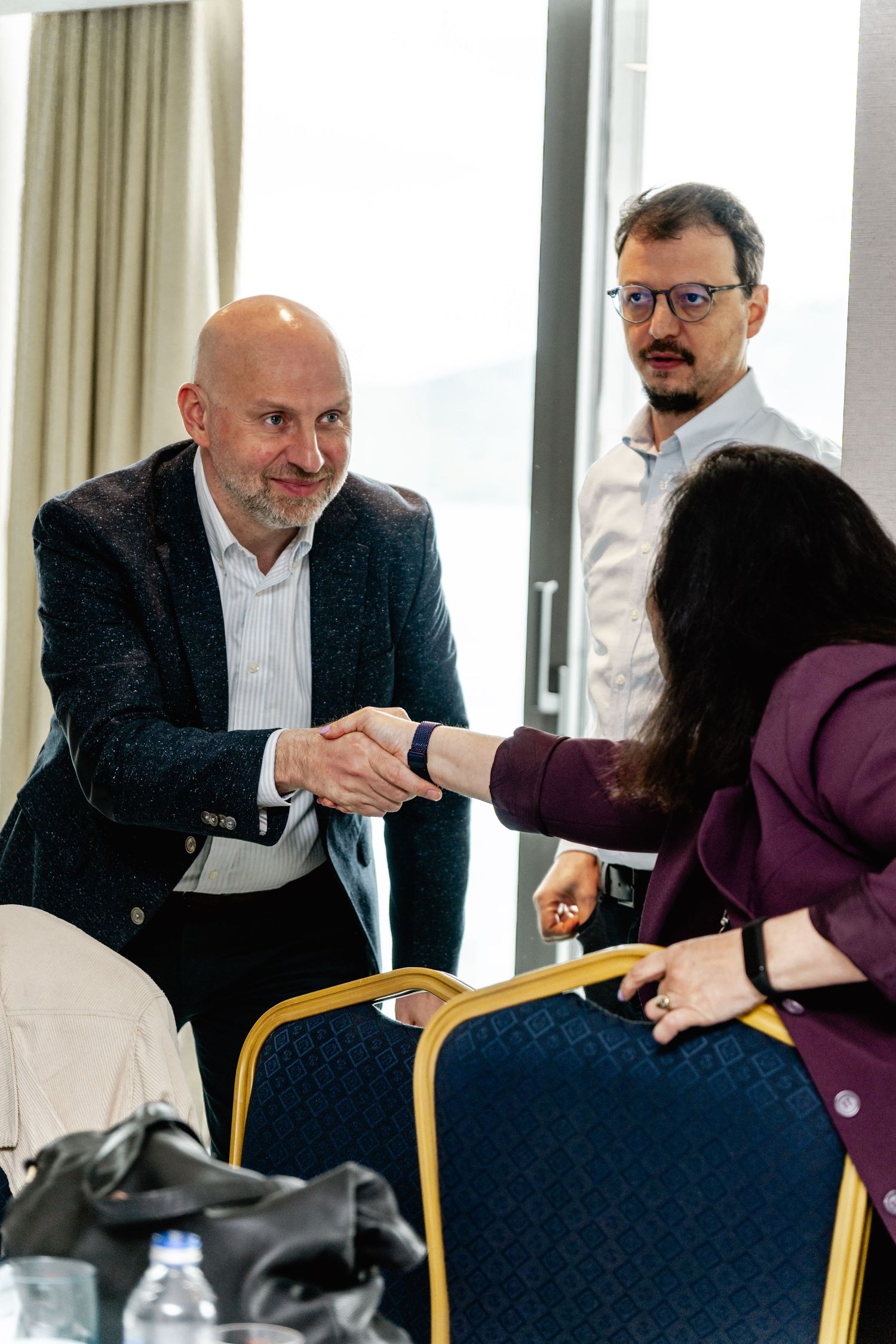 Bald businessman in blazer shaking hands with woman in purple jacket while colleague watches in modern conference room.