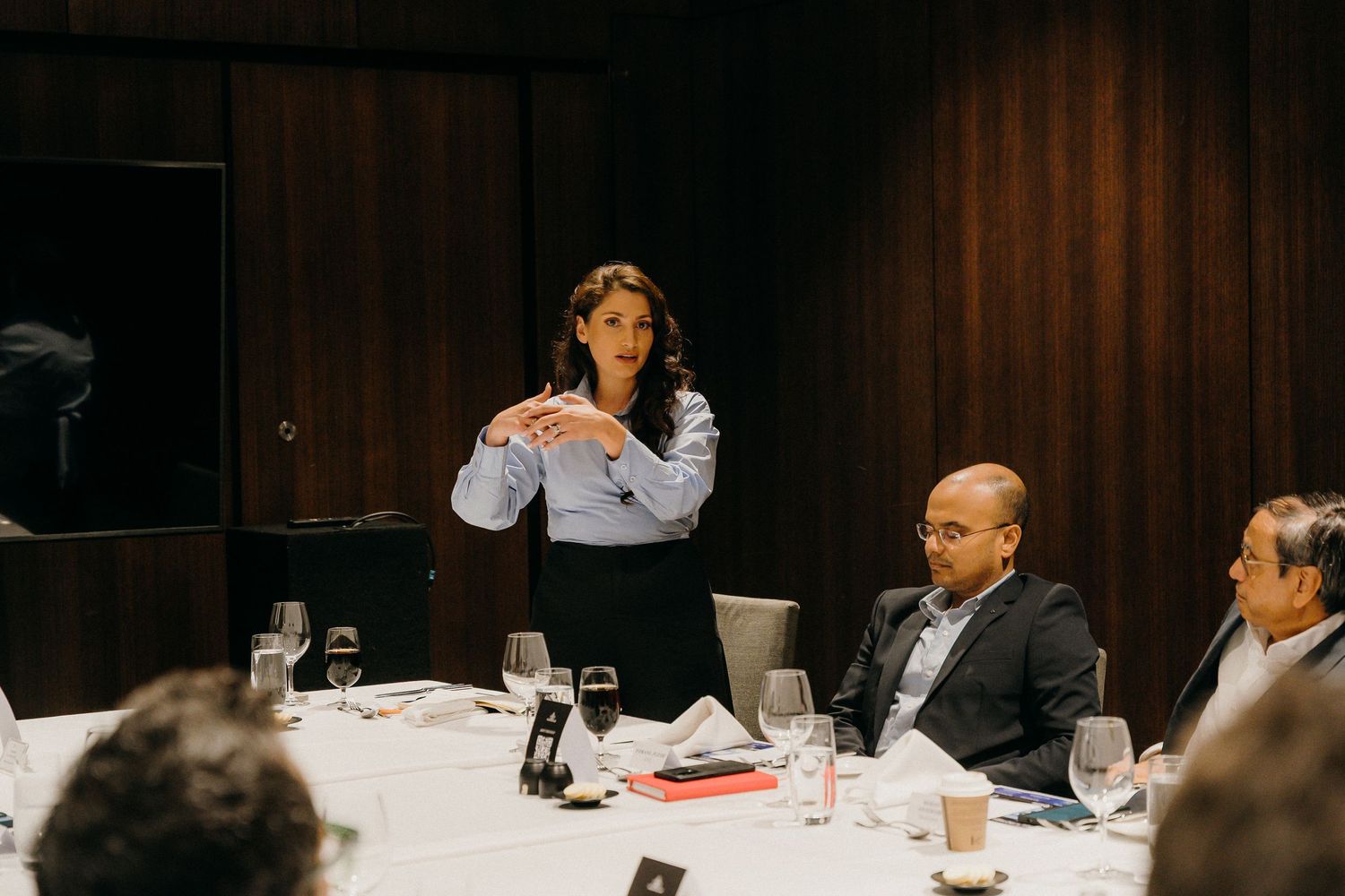 Professional woman presenting at a business dinner meeting with colleagues seated around a formal table.