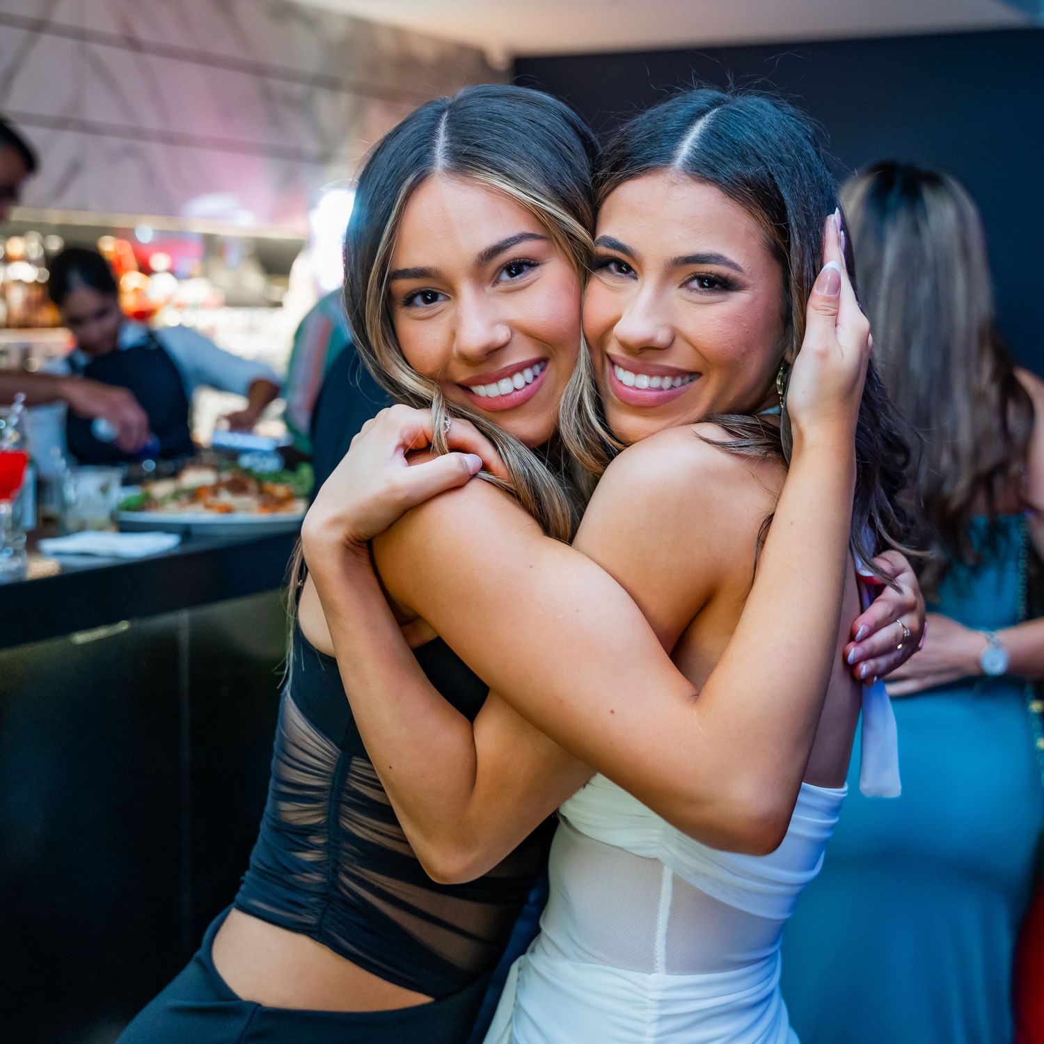 Two friends embrace and smile while enjoying a night out at a bar with dim lighting in the background.